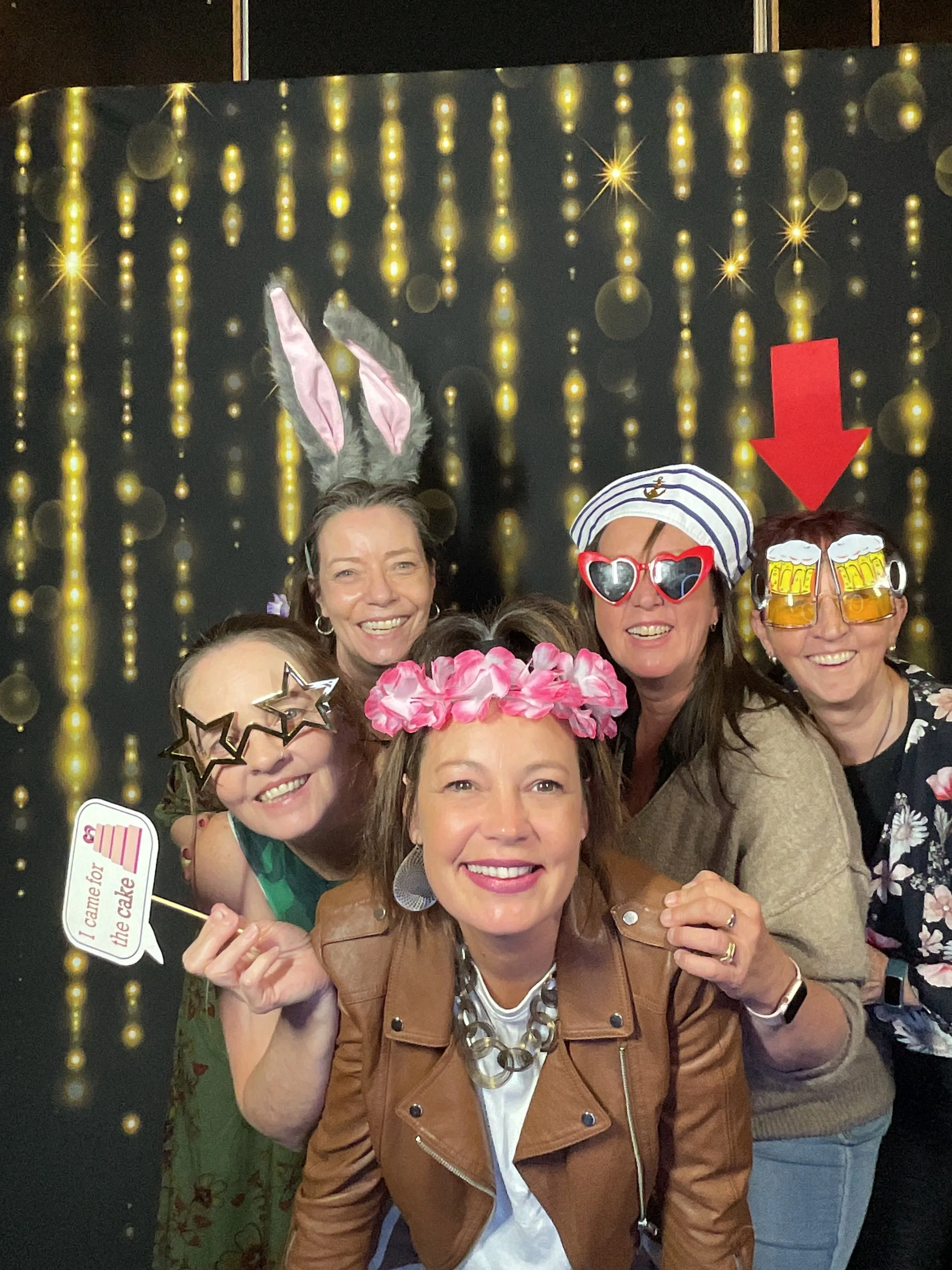 Group of five women celebrating, wearing fun accessories like bunny ears, star-shaped glasses, a floral headband, a sailor hat, and beer glasses. They are smiling in front of a festive, sparkly backdrop with hanging lights.
