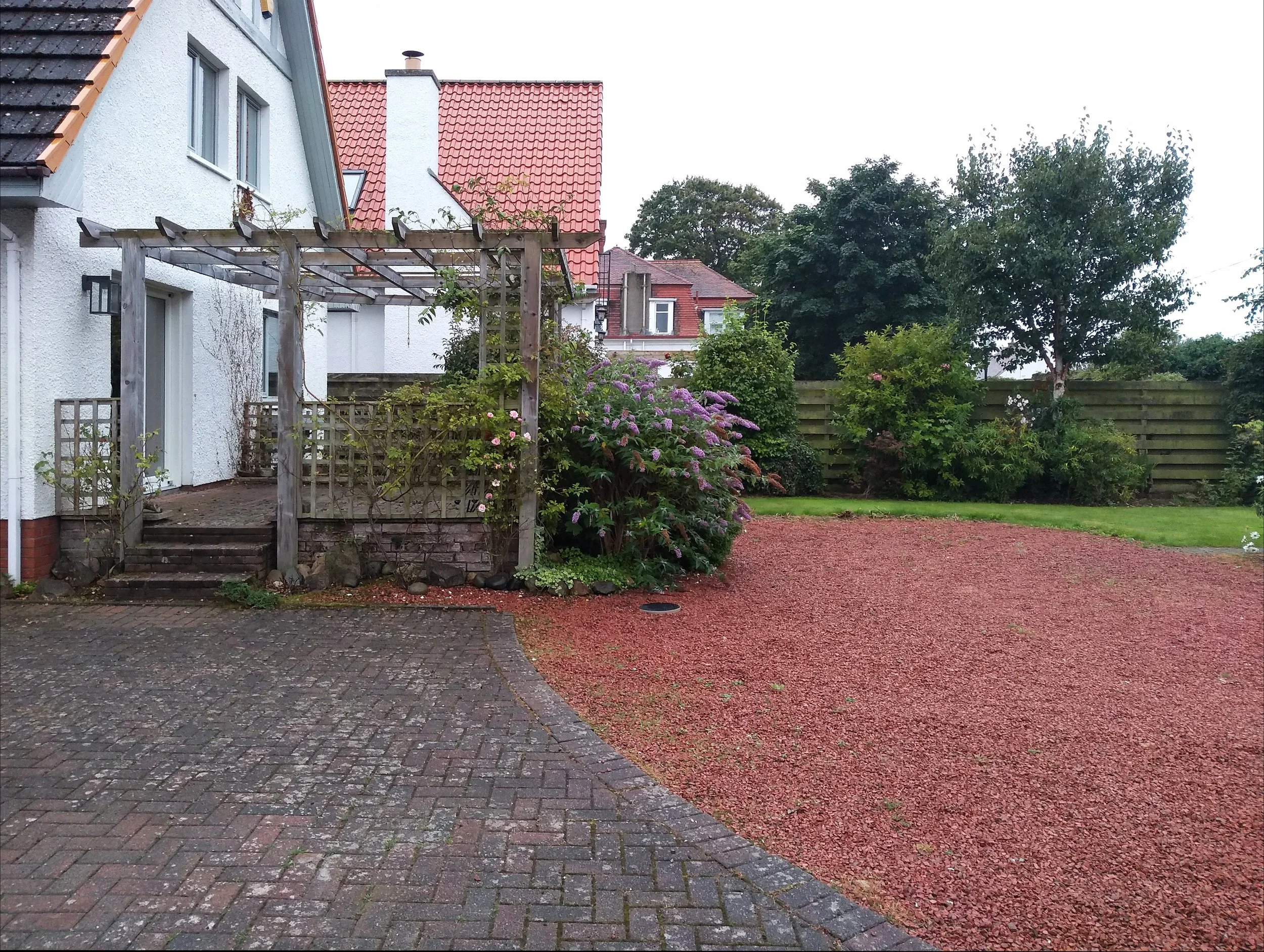 A pergola inf ront of a white house, with a paved parking area and red gravel surrounding it.