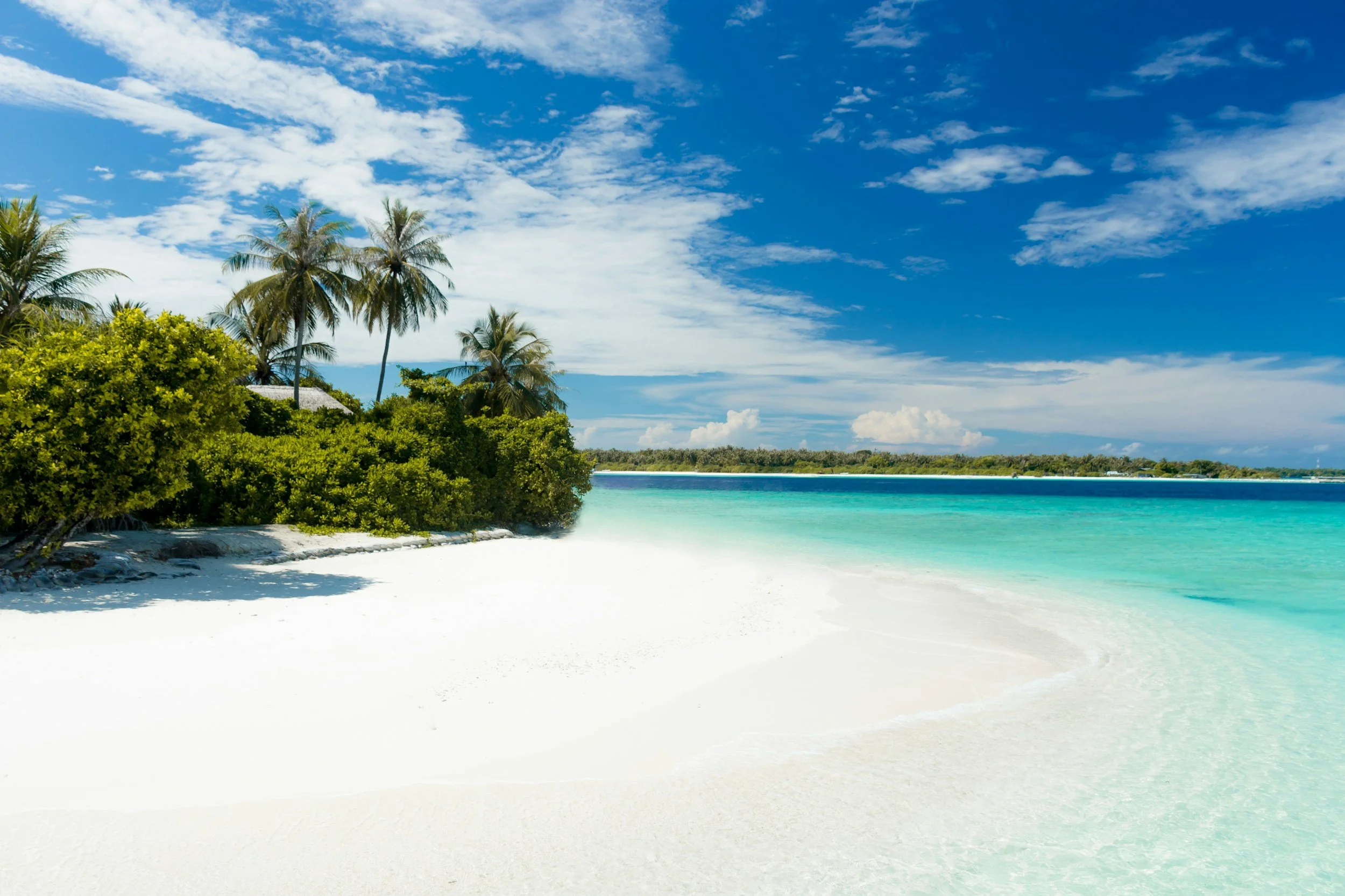 Tropical beach with white sand, turquoise water, green bushes, and palm trees under a partly cloudy blue sky.