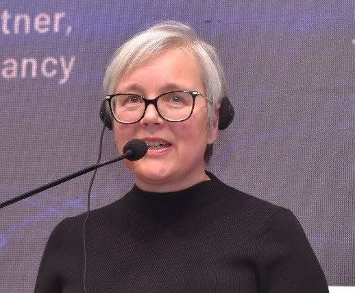 Caroline Bremner wearing headset with a microphone, speaking in front of a presentation screen.