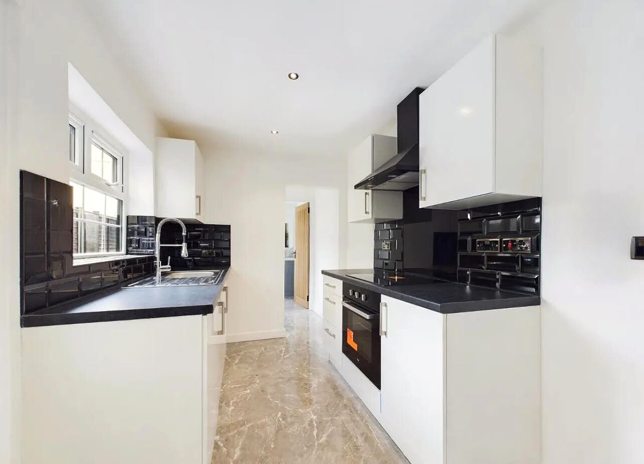 Modern kitchen with white cabinets, black countertops, and black tile backsplash. Features include a stainless steel oven, a stove, and a chrome faucet over the sink. Bright, minimalist design with marble flooring.