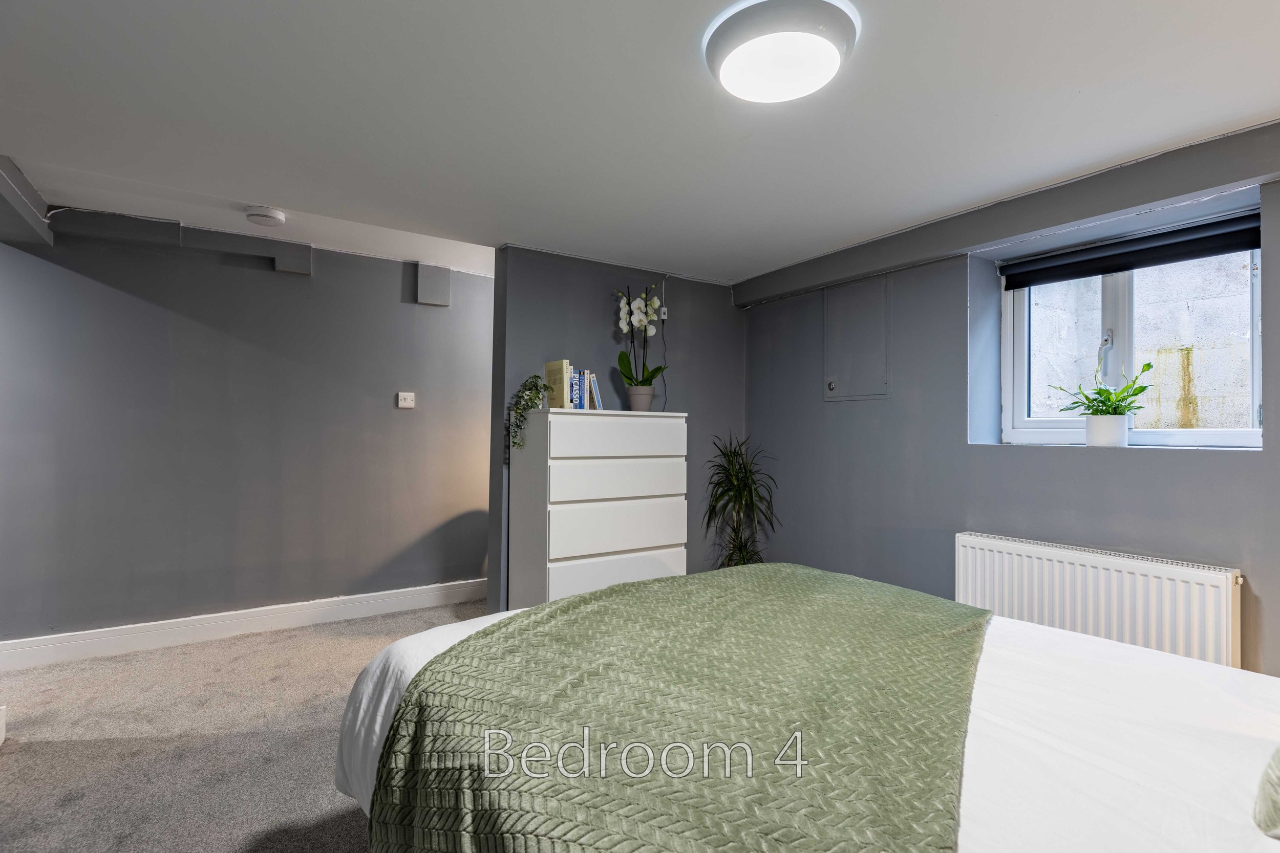 Bedroom interior with gray walls, a white chest of drawers, a green blanket on the bed, a small window with a potted plant, and a ceiling light.