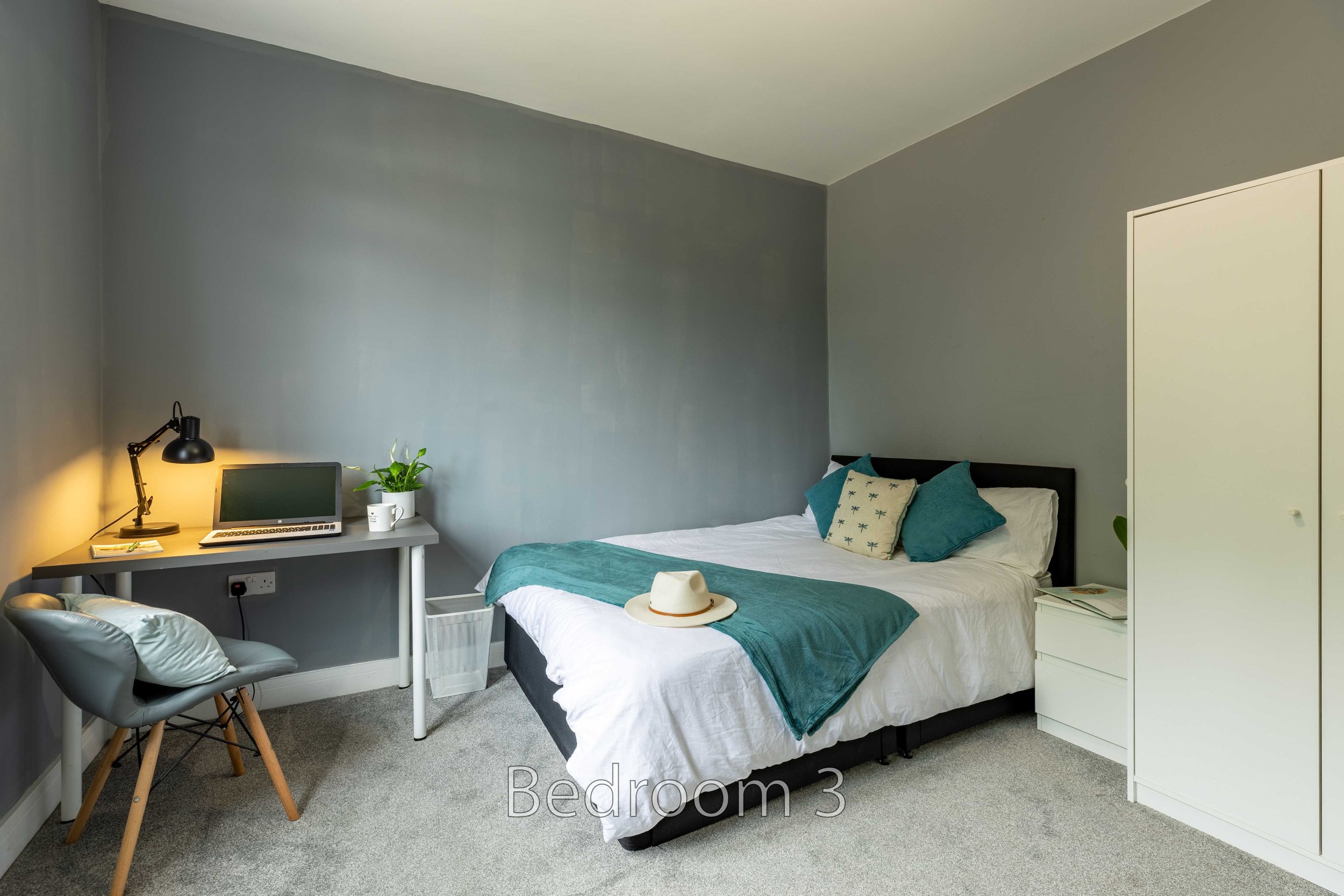 Modern bedroom with gray walls, a bed with white and teal bedding, a desk with a laptop and lamp, and a white wardrobe.