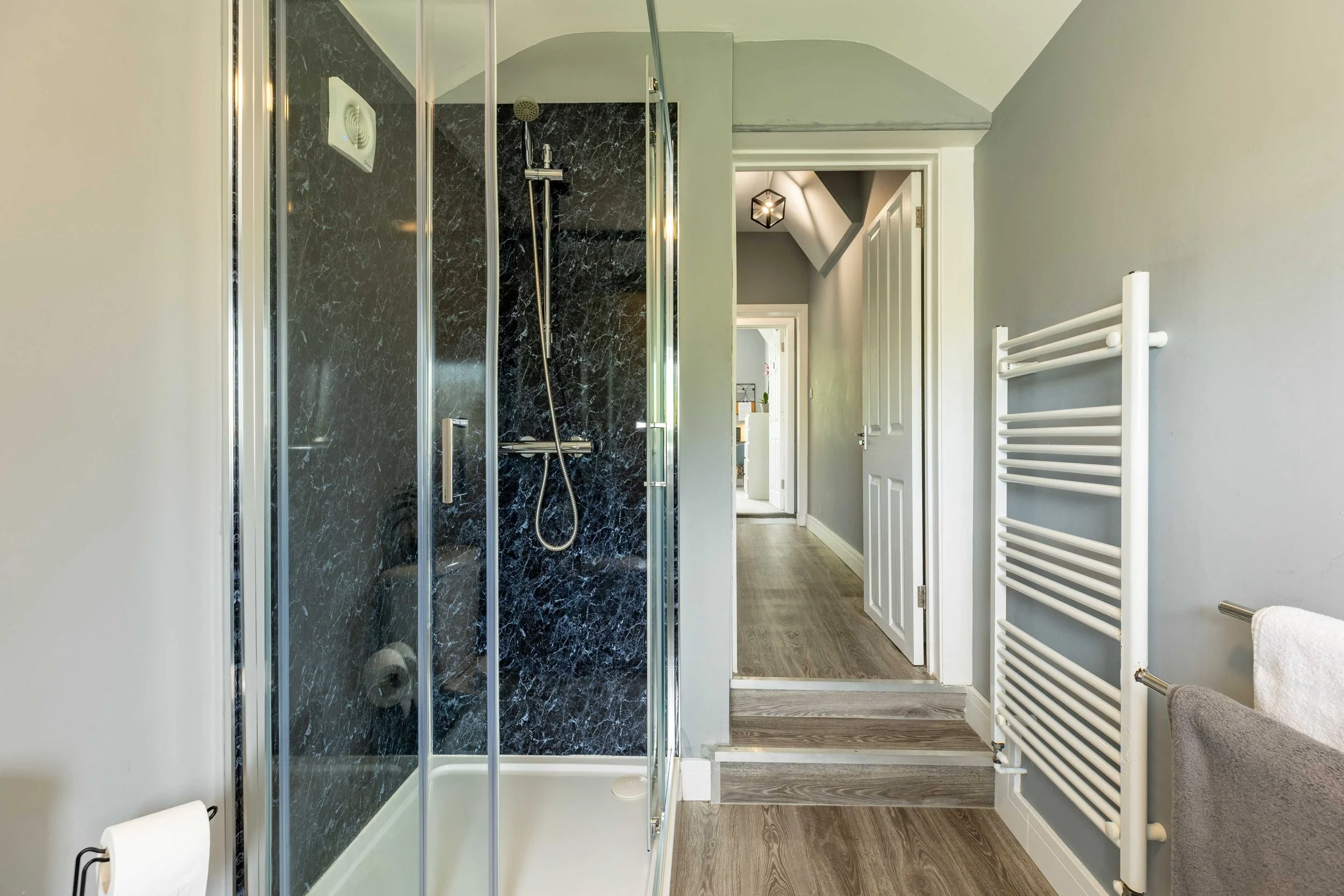 Modern bathroom with glass shower enclosure, gray walls, and a heated towel rail, connected to a hallway with wooden flooring and white doors.