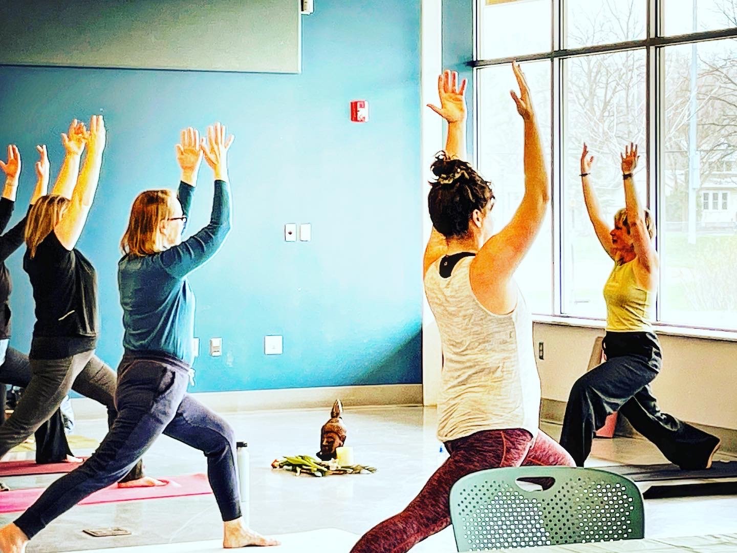 Yoga class in session with participants practicing warrior pose in a bright room with blue walls and large windows.