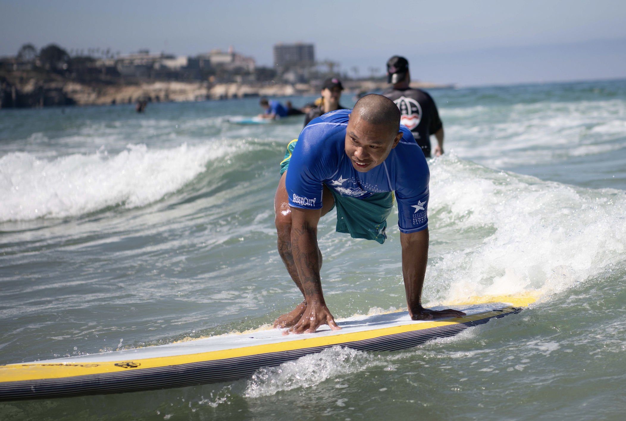 Silhouette of a surfer walking from the ocean towards the beach, with people in the background and palm trees visible on the shore.