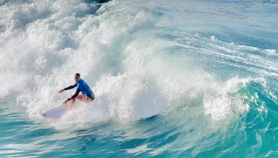 Surfer riding a wave in the ocean wearing a blue wetsuit, with whitewater and turquoise water surrounding them.
