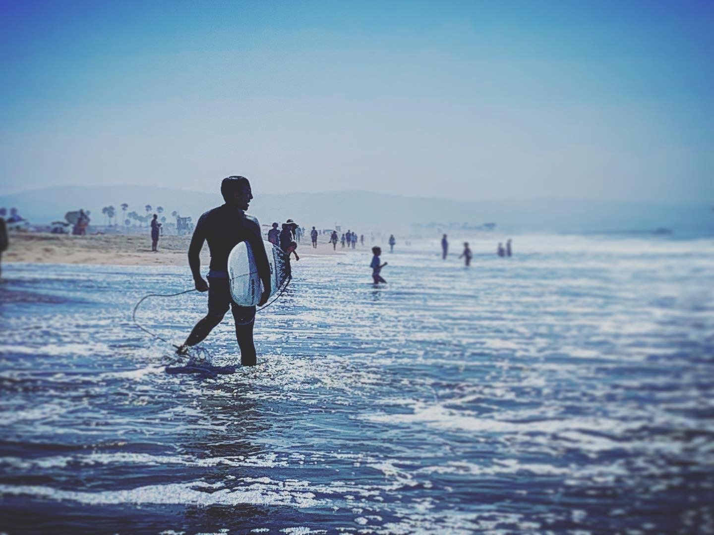 Surfer walking on a beach with a surfboard, ocean waves in the foreground and people in the distance. It's a sunny day.