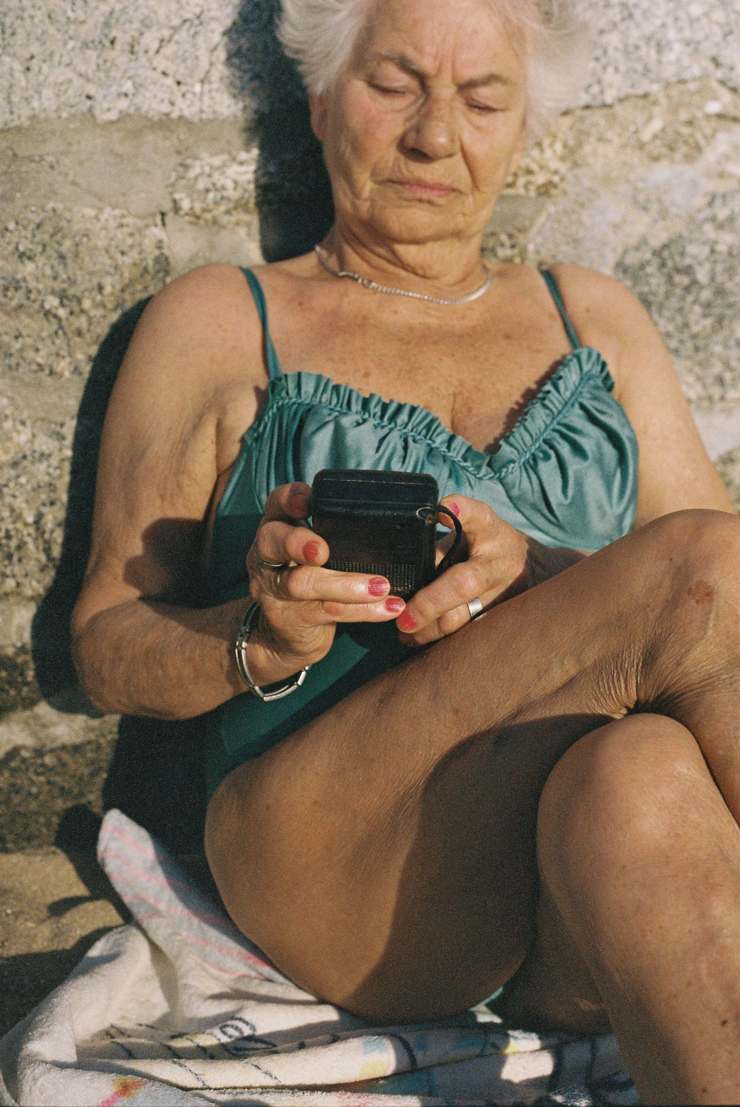 Elderly lady sunbathing at the beach holding retro radio in hand.