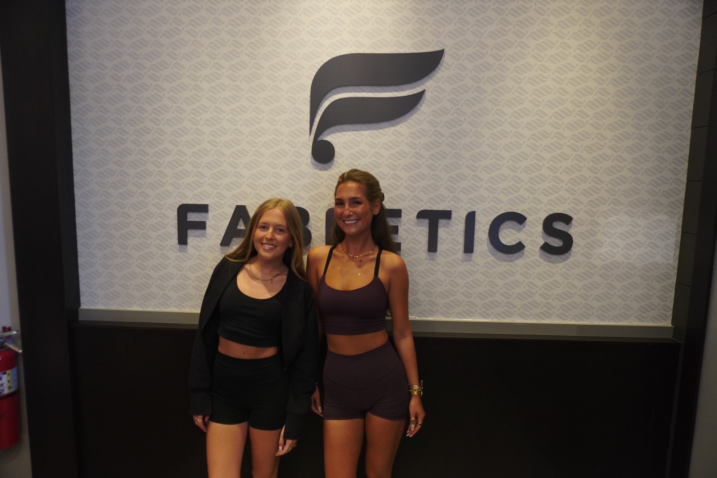 Two young women smiling and standing in front of a sign that reads "Fabletics" with a logo above it. One woman has long blonde hair and is wearing a black crop top and black shorts, while the other has long brown hair and is wearing a purple sports bra and matching shorts.