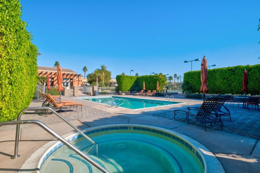 Outdoor pool area with hot tub, lounge chairs, umbrellas, and surrounding foliage under a clear blue sky.