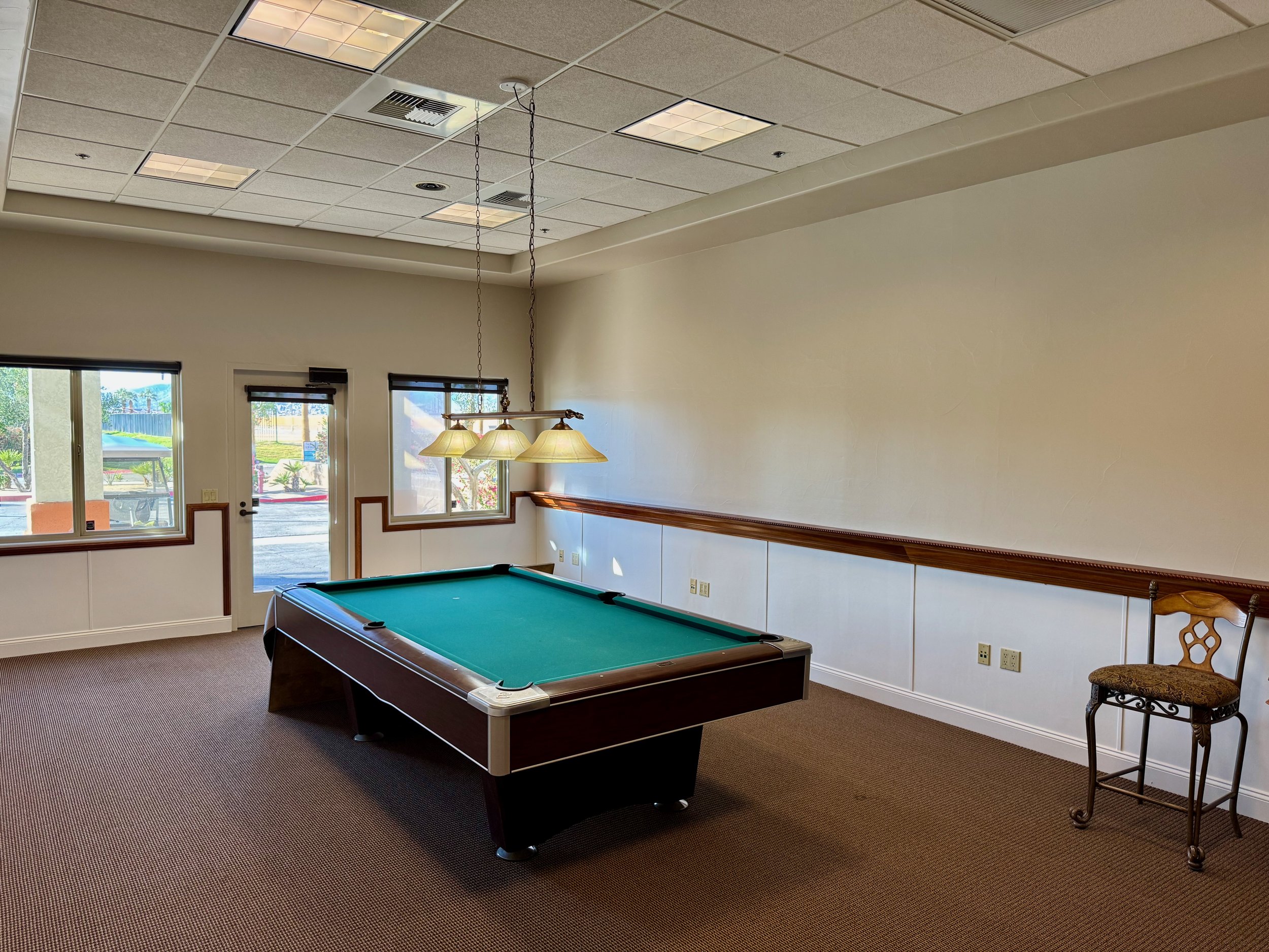 Room with pool table and high chair, large windows, ceiling lights, and beige walls.