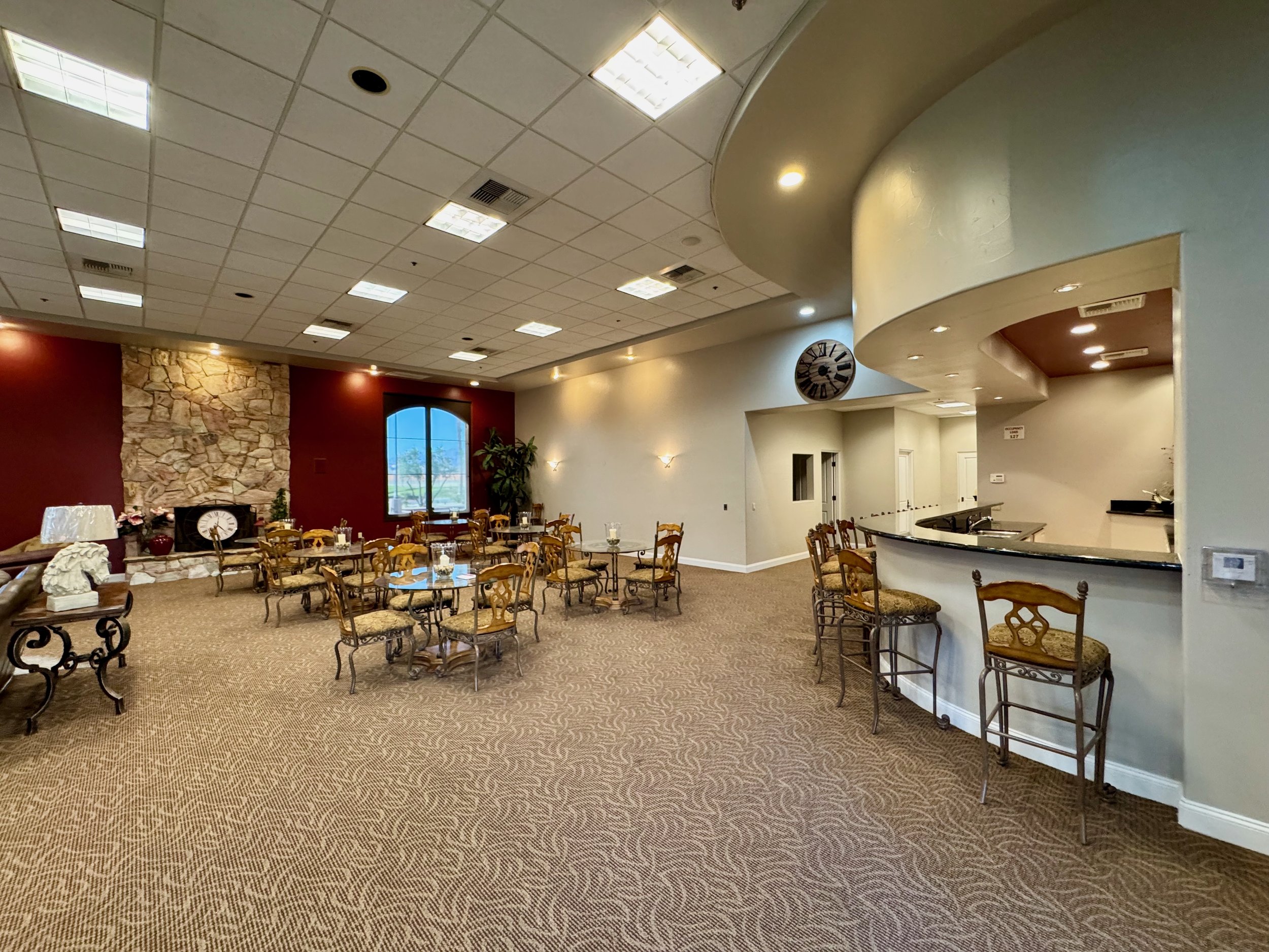 Interior of a spacious lounge area with tables and chairs, a stone fireplace, maroon walls, a semi-circular bar, and a large window. The ceiling is fitted with fluorescent lights.