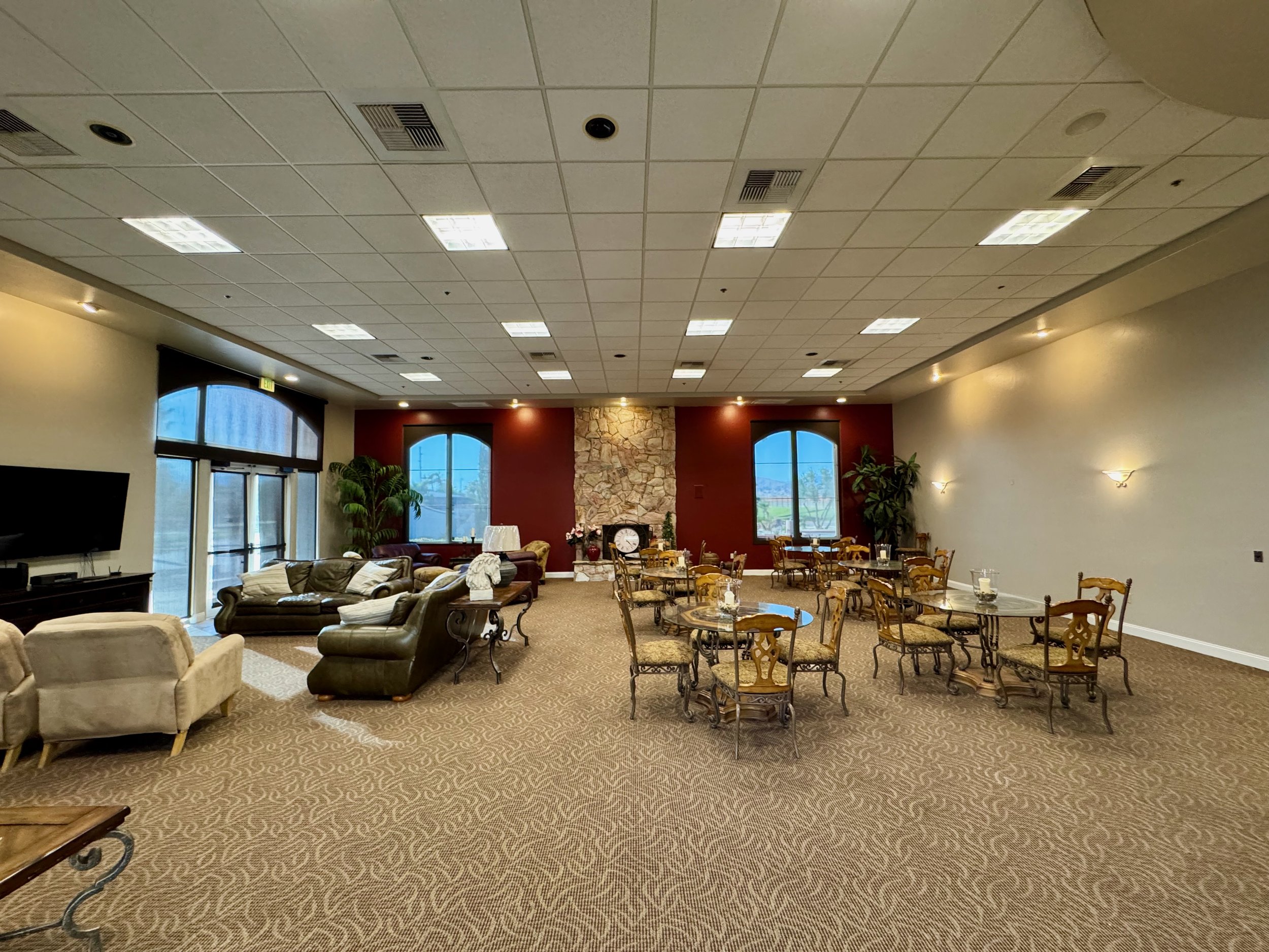 Spacious interior room with beige carpet, sofas, armchairs, small round tables with chairs, stone wall, large windows, and plants.