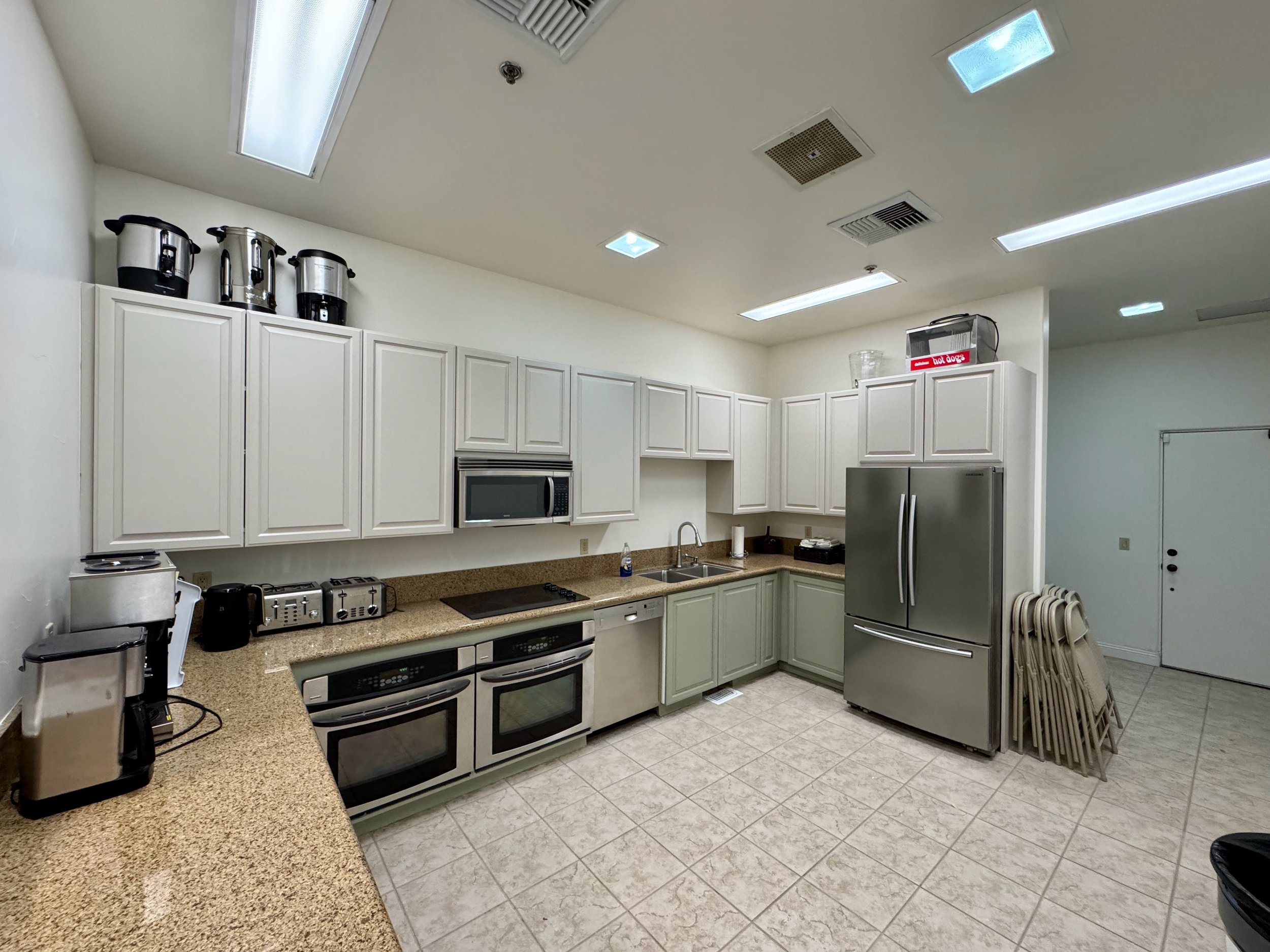 Modern kitchen with white cabinets and stainless steel appliances, including a double oven, refrigerator, microwave, and multiple toasters. Countertops feature various kitchen appliances, such as coffee makers and slow cookers. The floor has beige ti