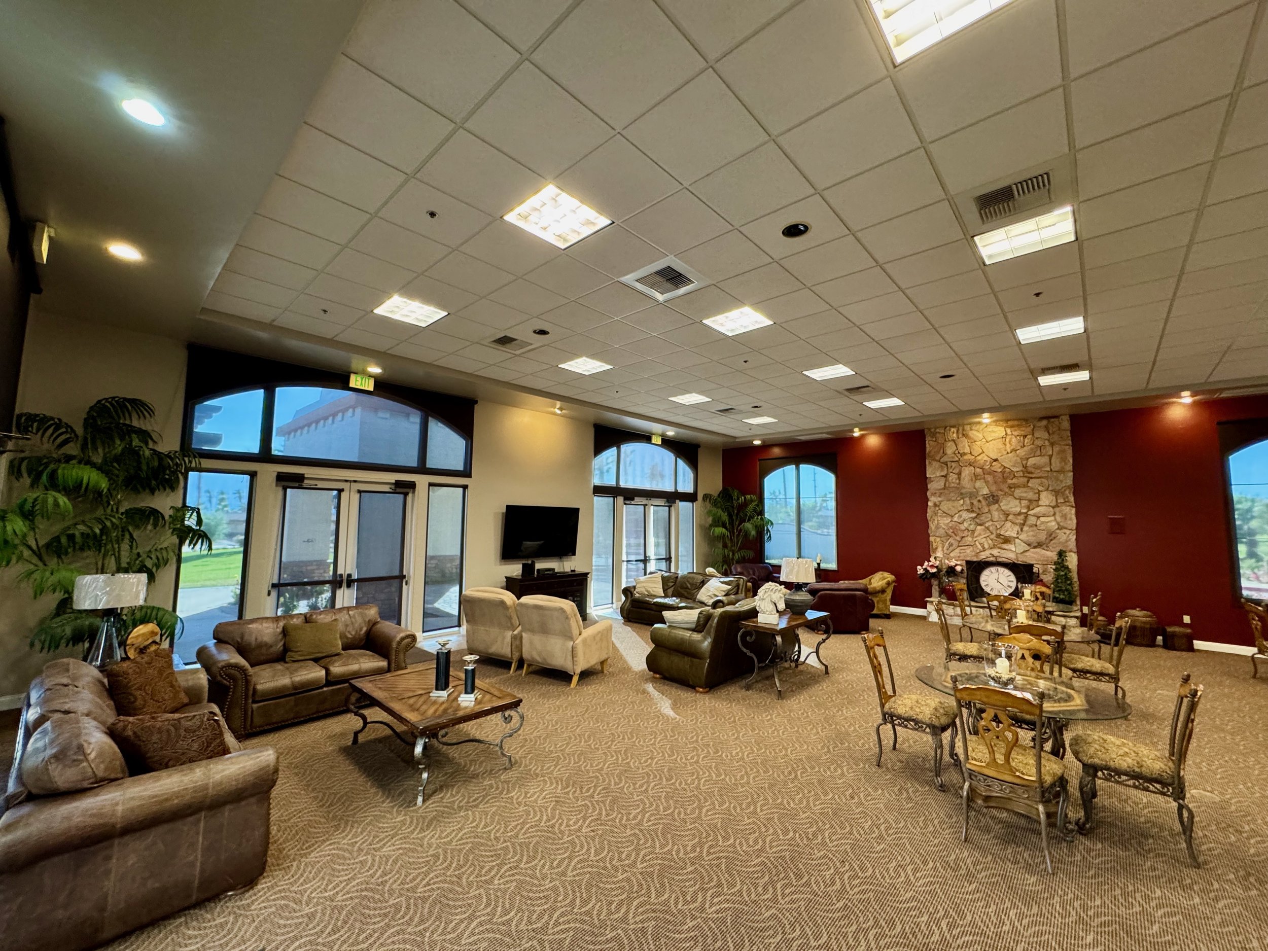 Spacious living room with multiple seating areas, featuring couches and armchairs. Glass tables with ornate chairs are set in the room. Large windows provide ample natural light. A stone accent wall with a fireplace and a large clock is visible. Indo