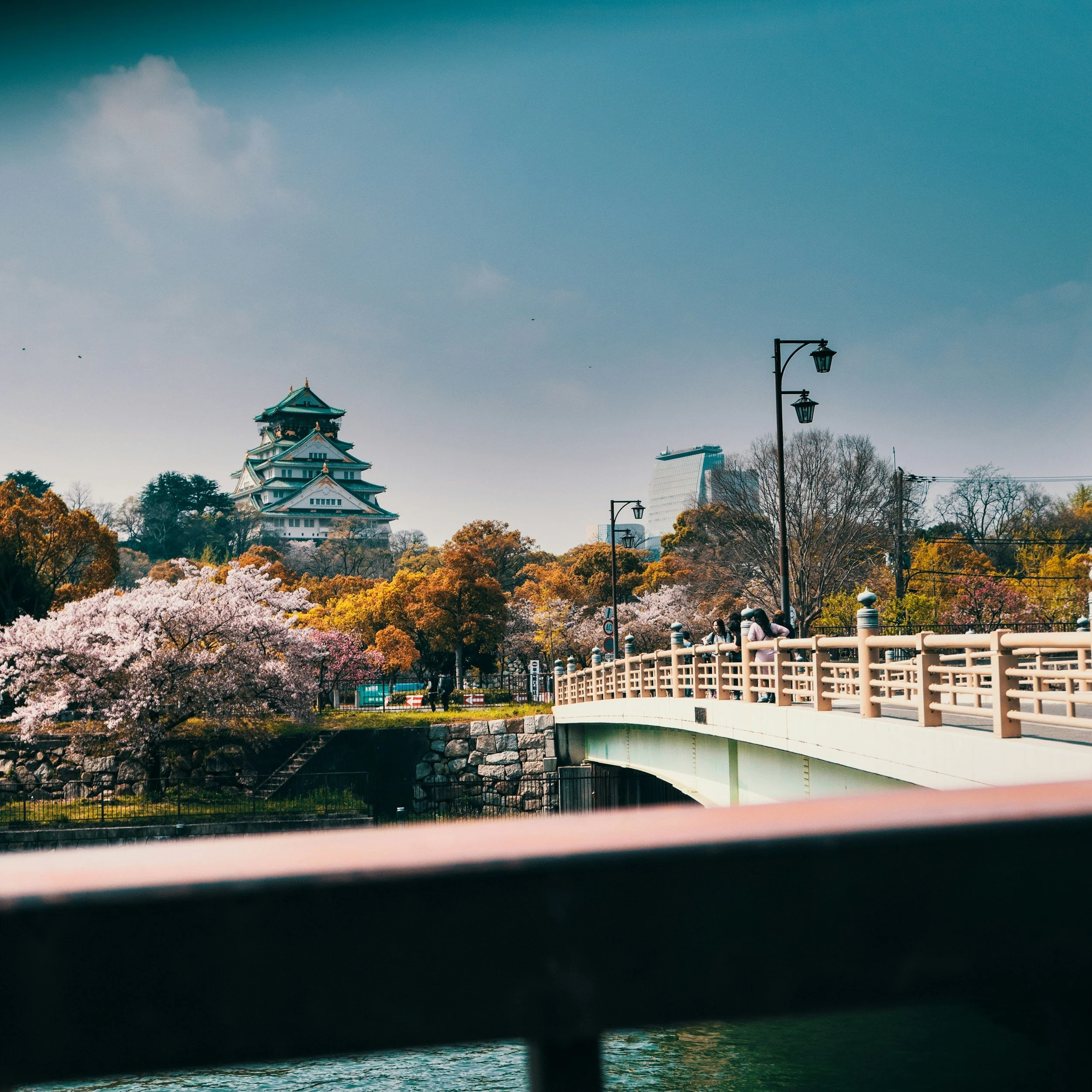 A scenic view of a historic Japanese castle with a multi-tiered roof, surrounded by flowering sakura trees and autumn-colored foliage, with a bridge and people walking across it in the foreground.