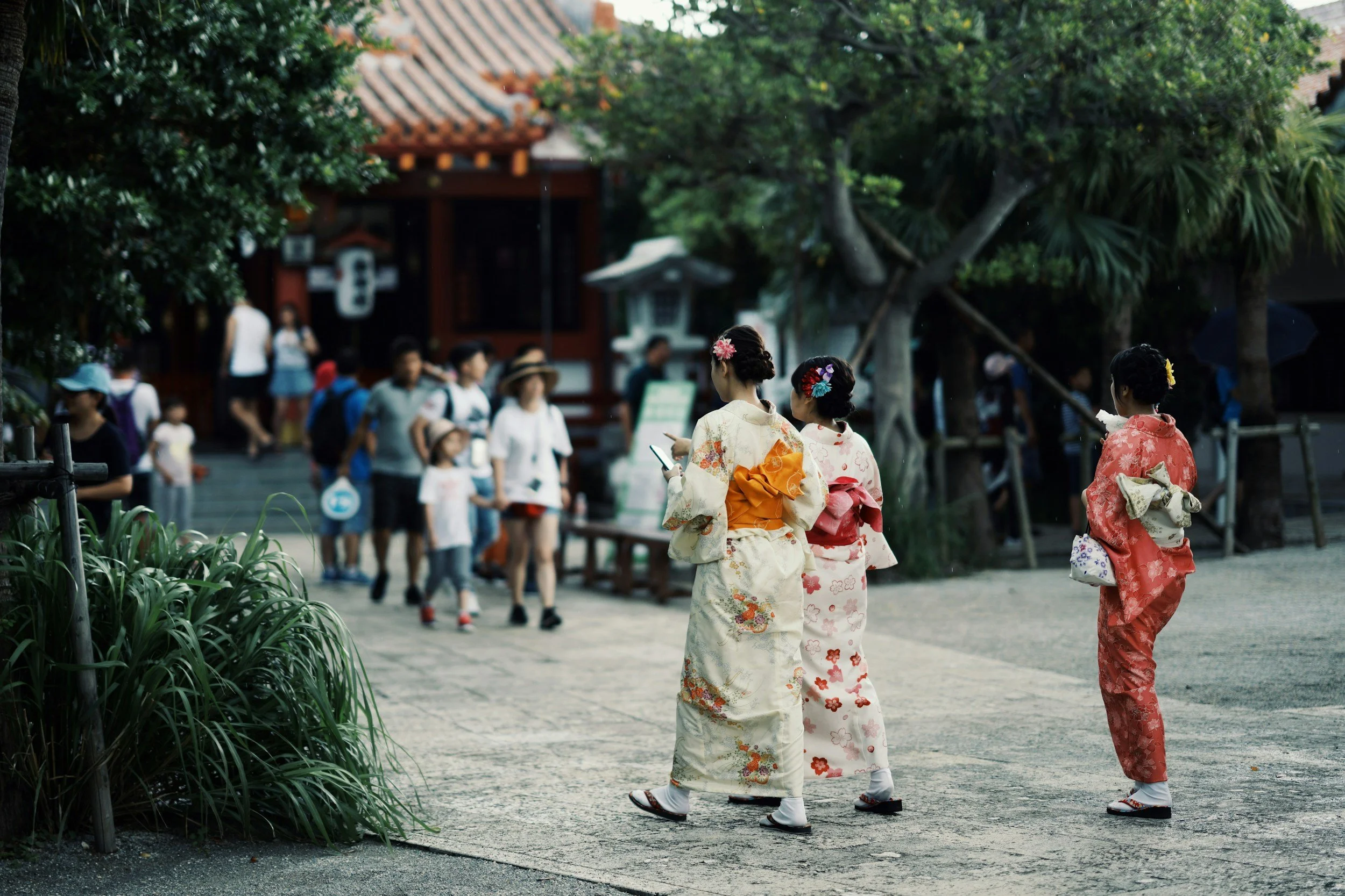 Three women in traditional kimonos walking in a park with a crowd of people and a temple in the background.
