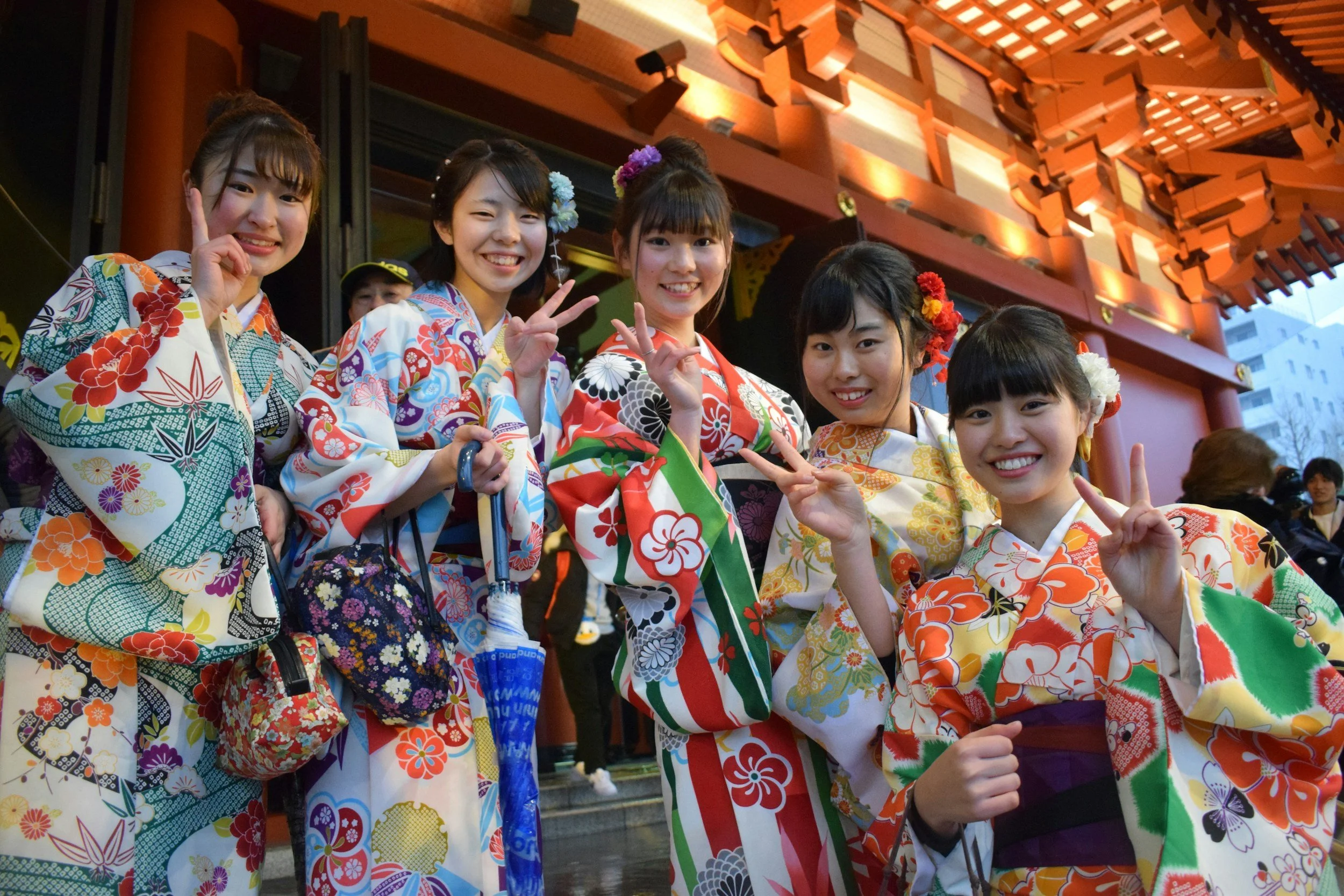 Six young women dressed in colorful traditional Japanese yukatas, smiling and making peace signs with their hands, standing outside a shrine with ornate wooden carvings.