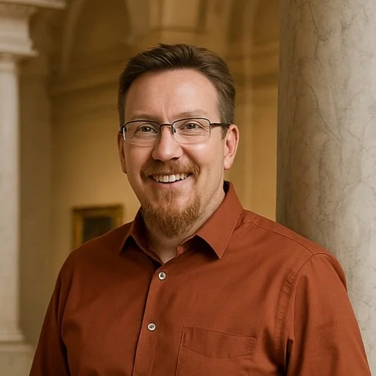 Smiling man with glasses and a beard, wearing a brown shirt, standing in a building with classical architecture.