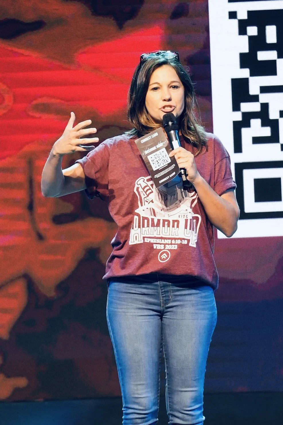 A woman with shoulder-length brown hair wearing glasses on her head, a maroon T-shirt, and jeans, speaking into a microphone on stage during a conference or event.