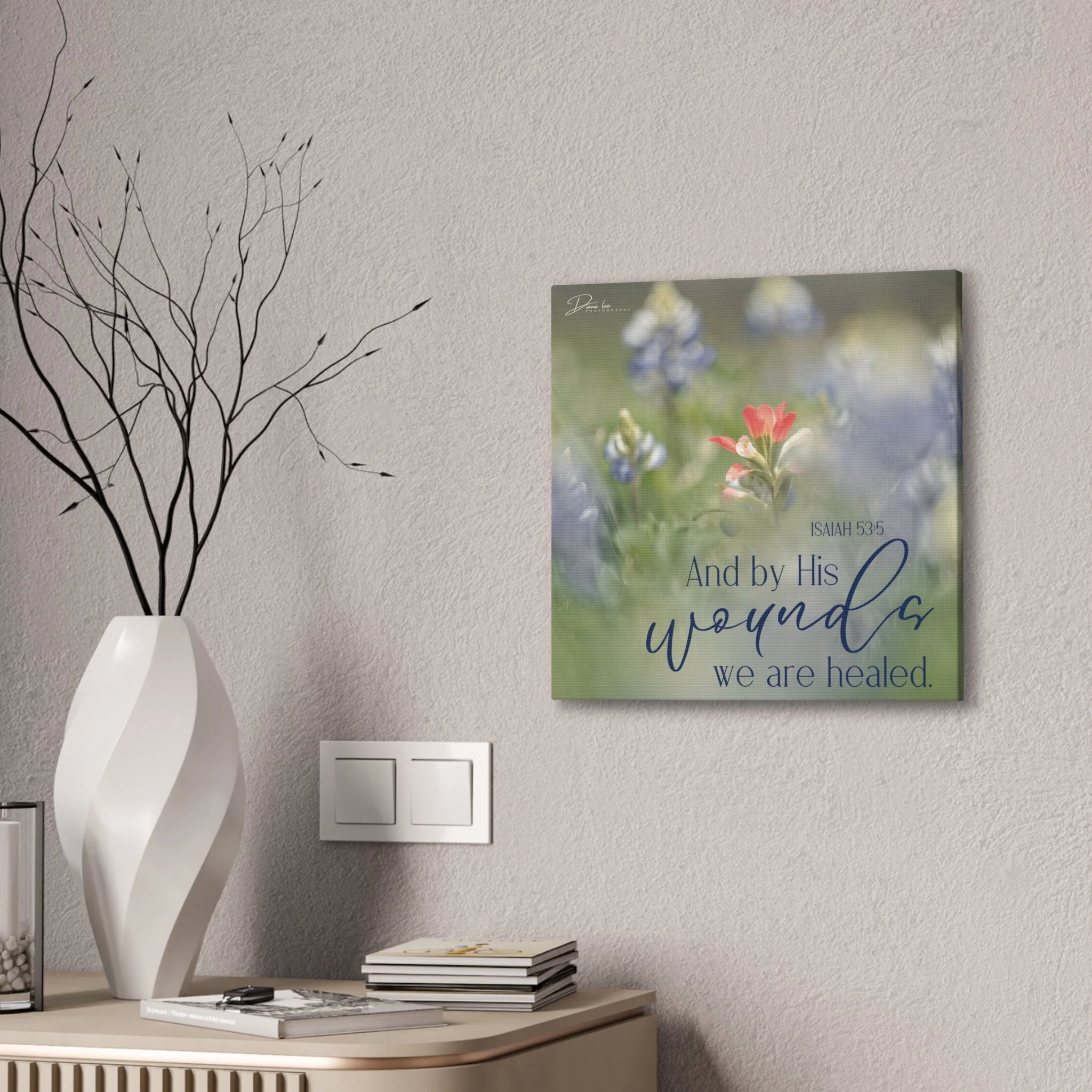A wall featuring a framed floral artwork with a Bible verse from Isaiah 53:5, and a white decorative vase with black twigs on a wooden side table.