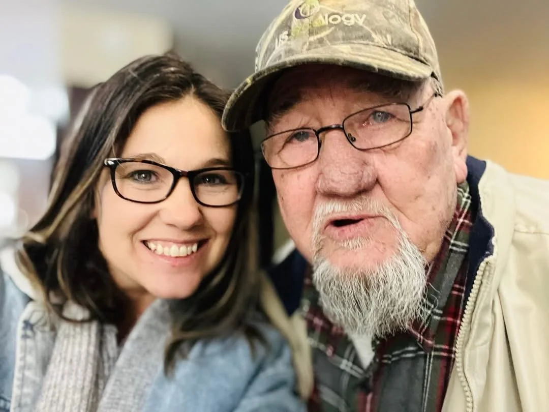A smiling woman with glasses and long dark hair taking a selfie with an older man with glasses, a gray beard, and a camouflage cap, both facing the camera closely.