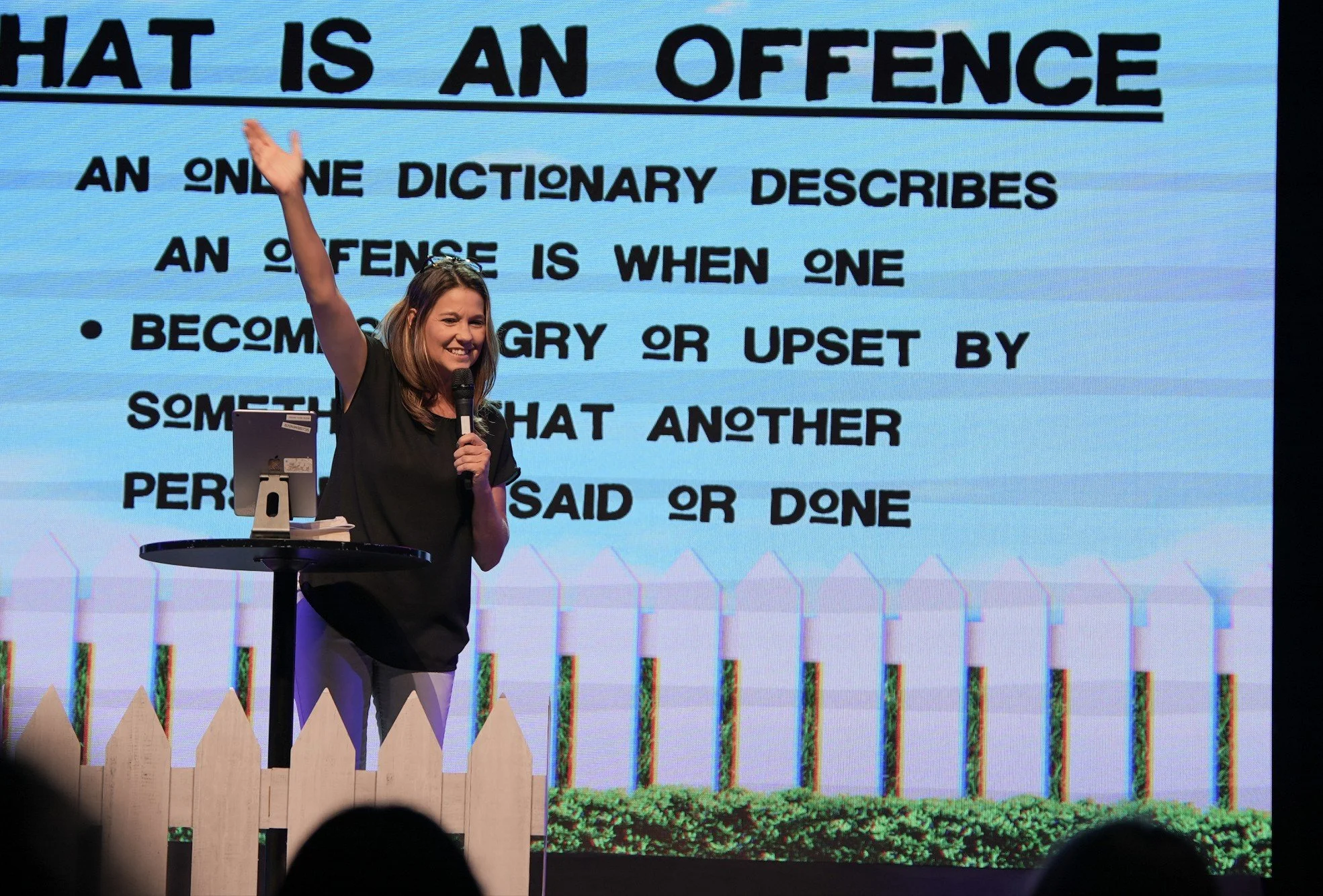 A woman with shoulder-length brown hair, smiling, holding a microphone and raising her left hand, standing on stage with a large screen behind her displaying a definition of offense.