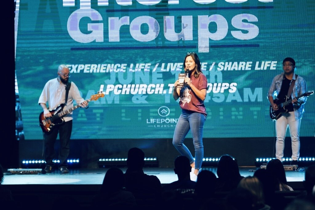 A woman singing on stage with two men playing guitars, in front of a large screen displaying the words 'Groups' and 'Experience life, live, share life' at a church event.