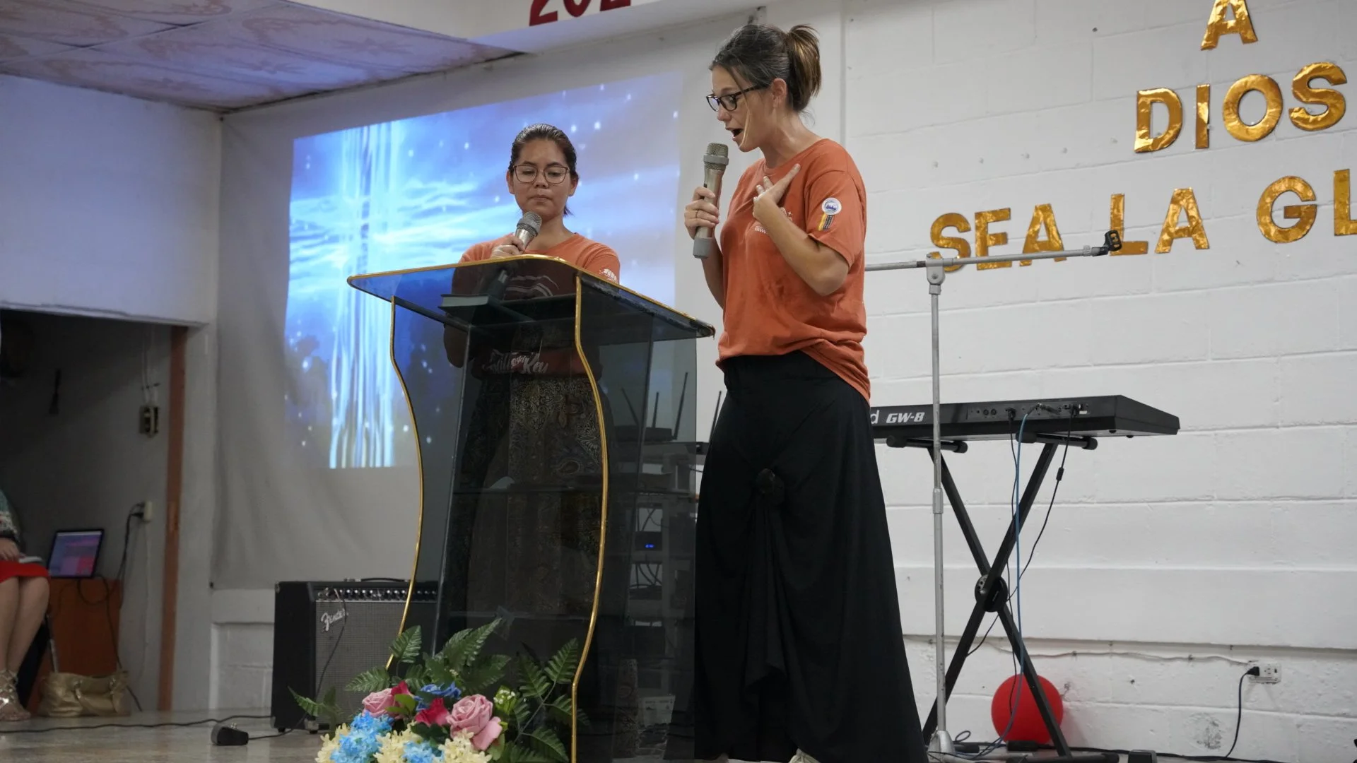 Two women standing at a pulpit, speaking into microphones during a presentation or event. One woman is reading from a paper, the other is gesturing with her hand. There is a music keyboard and a projector screen in the background, with decorative gold letters on the wall that read 'Sea la Gl...' indicating part of a larger phrase, and a bouquet of pink, blue, and white flowers in front of the pulpit.