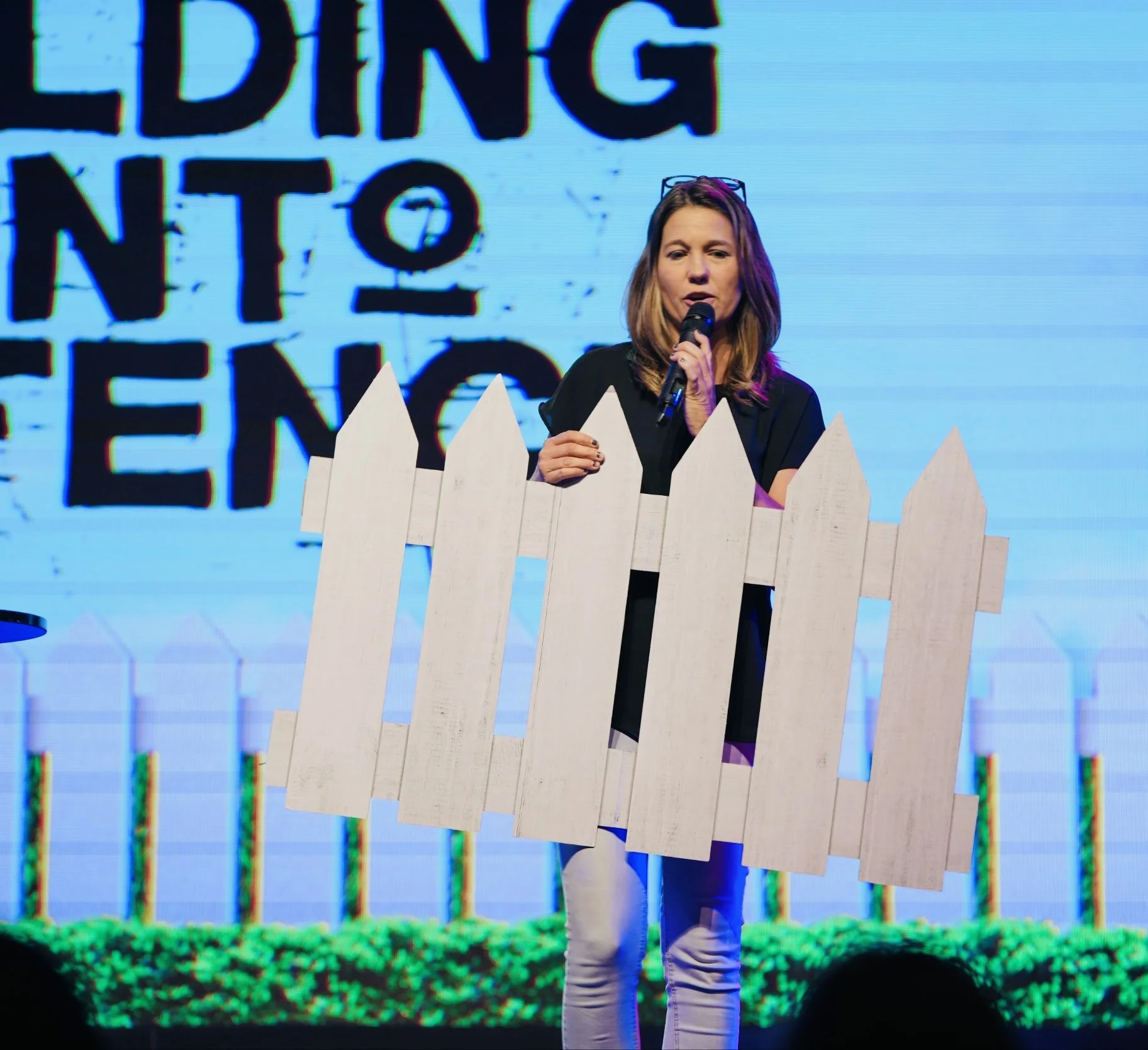 Woman speaking into microphone behind a white picket fence on stage with a colorful background.