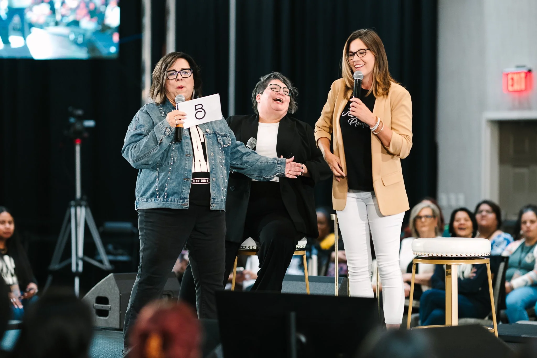 Three women on stage holding microphones, engaging with an audience during a conference or event, with seated attendees in the background.