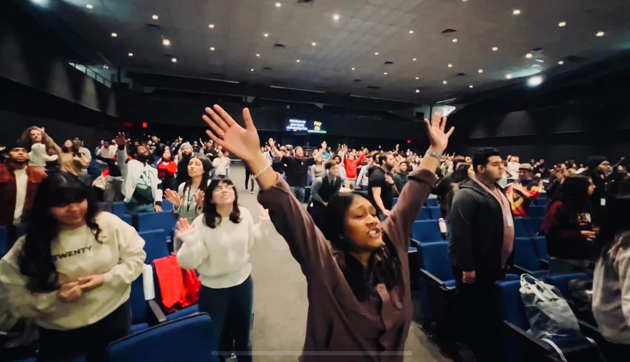 A large group of people standing in a dark auditorium with blue seats, some with hands raised, appearing engaged in a group activity or worship.
