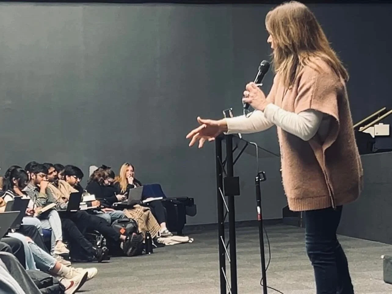 A woman speaking into a microphone in front of a seated audience, some taking notes or using laptops, in a classroom or conference room setting.