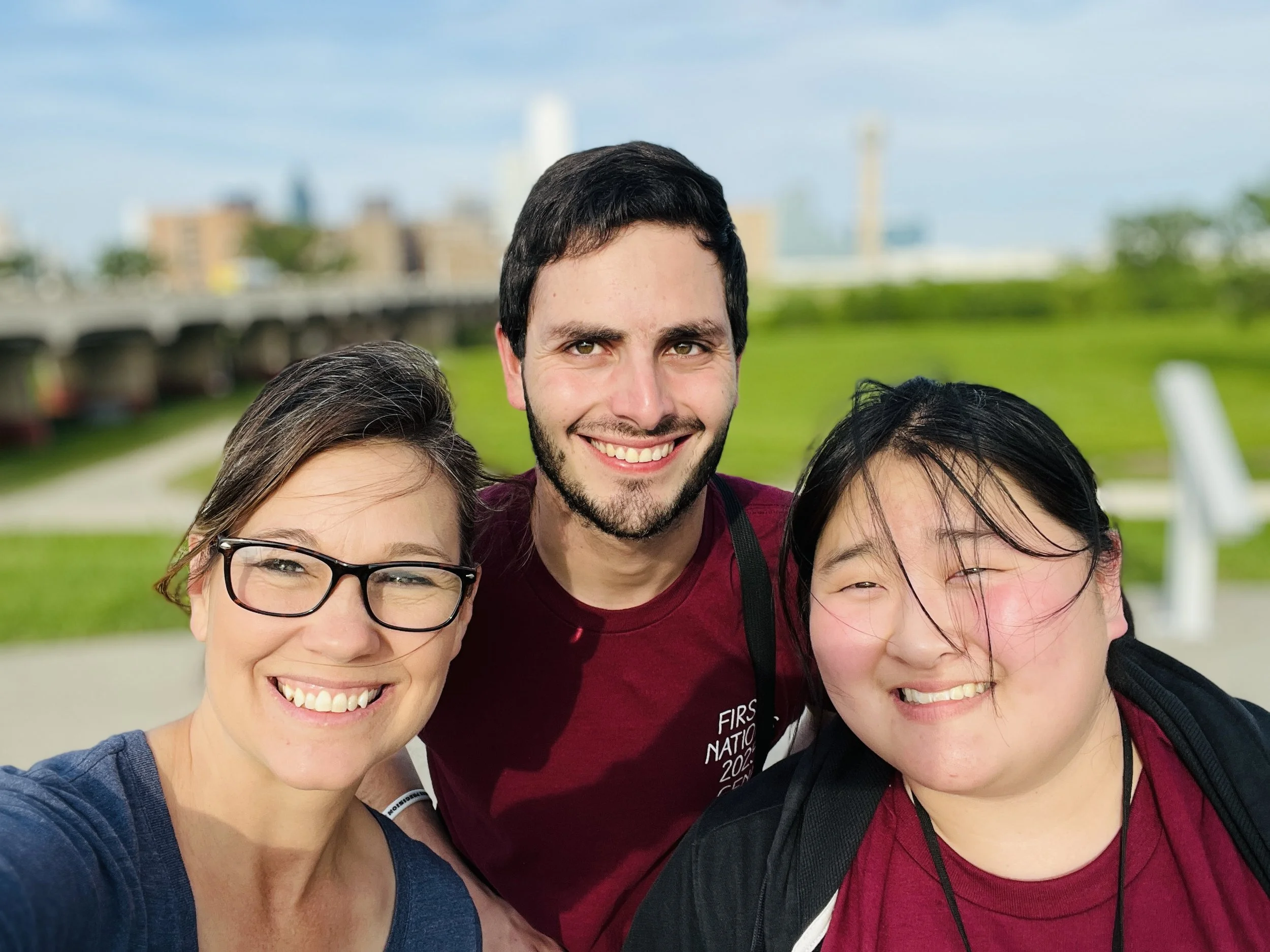 Three young adults taking a selfie outdoors in a park with a city skyline in the background. They are smiling, with the person on the left wearing glasses, the person in the middle with short dark hair and a beard, and the person on the right with long black hair.