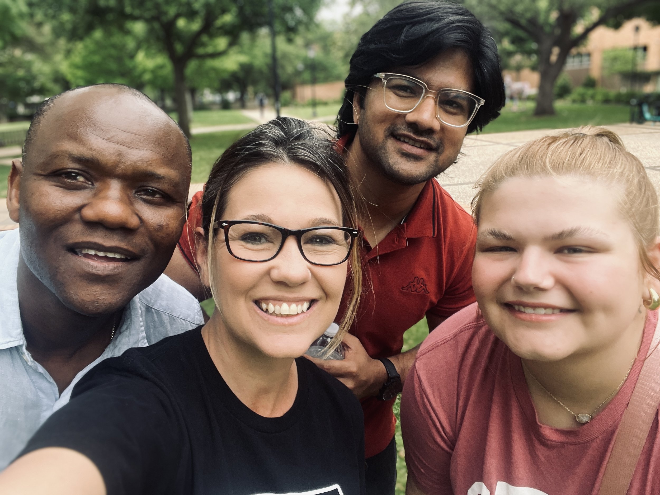 Group of five diverse people smiling for a selfie outdoors in a park with trees and a gazebo in the background.