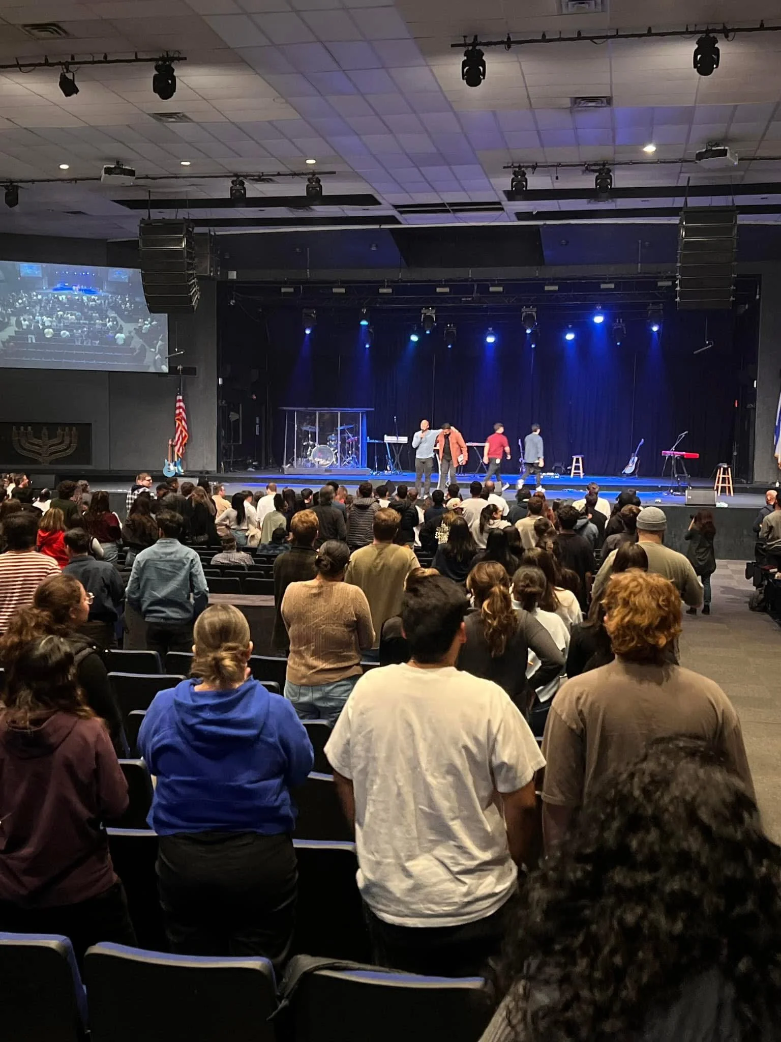 Audience standing and watching a stage with performers in a large auditorium, stage lighting illuminating the stage, and a drum set enclosed in a transparent box.