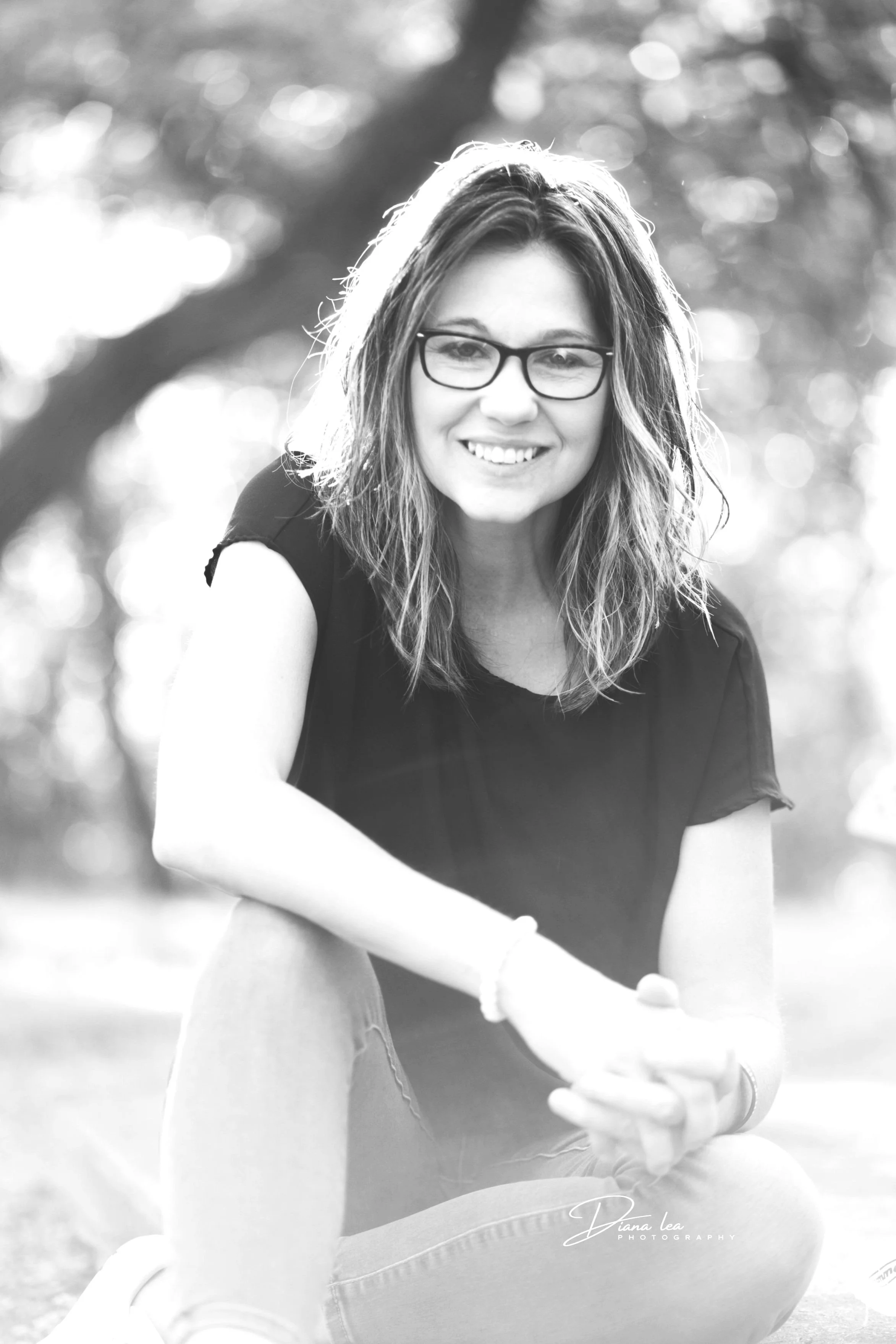 Black and white photo of a woman sitting outdoors, smiling, wearing glasses, a black t-shirt, and jeans, with trees in the background.