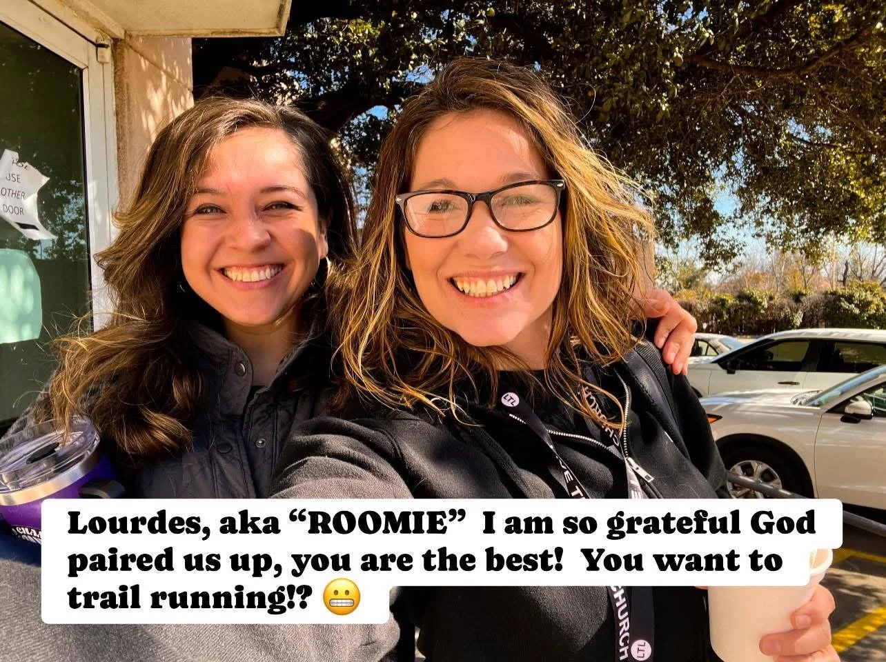 Two women smiling and taking a selfie outdoors in a parking lot with cars and trees behind them.