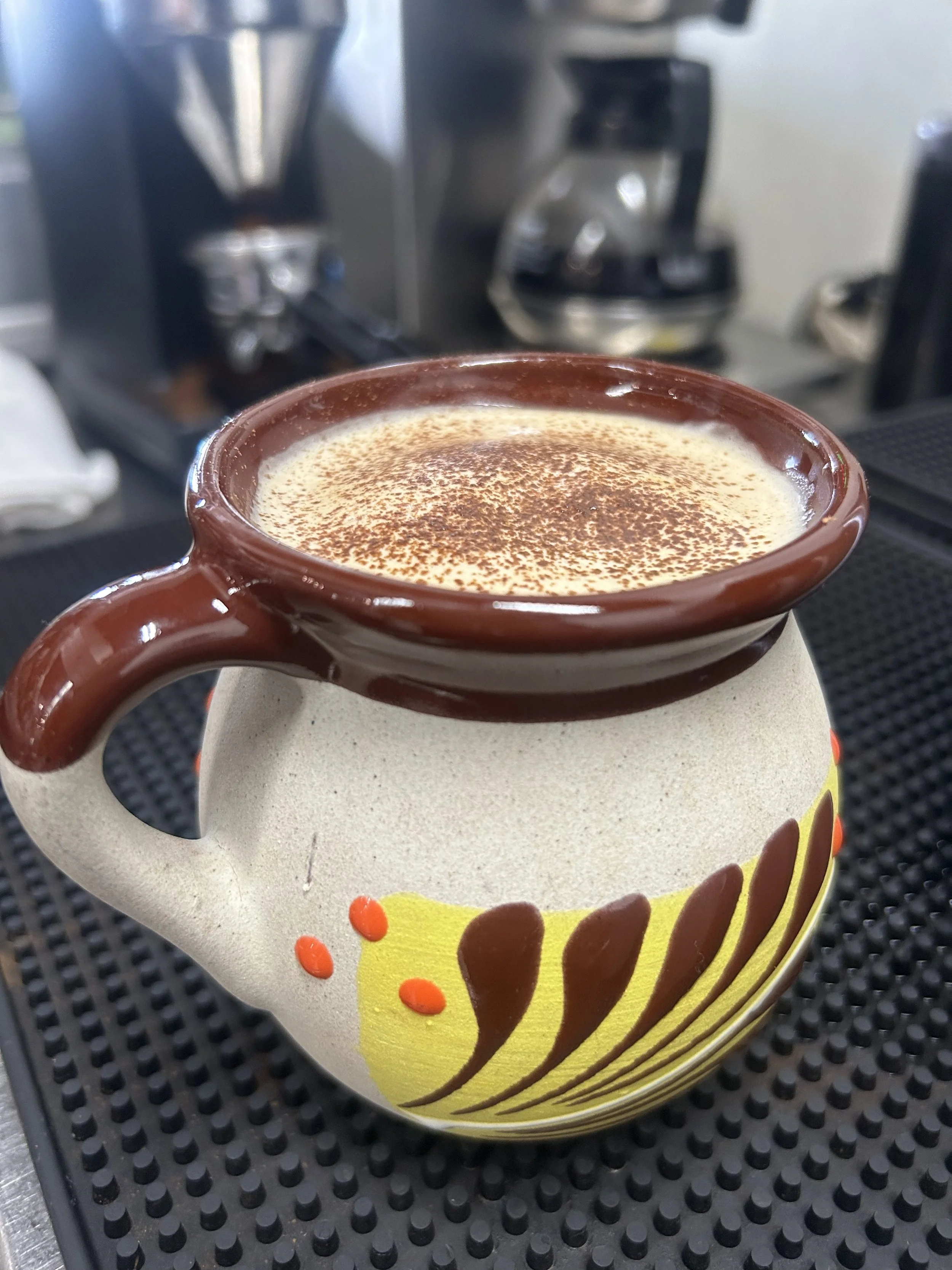 A ceramic pitcher filled with a creamy beverage topped with cocoa powder, with a brown handle and a decorative yellow and brown design on the body, placed on a black rubber mat.