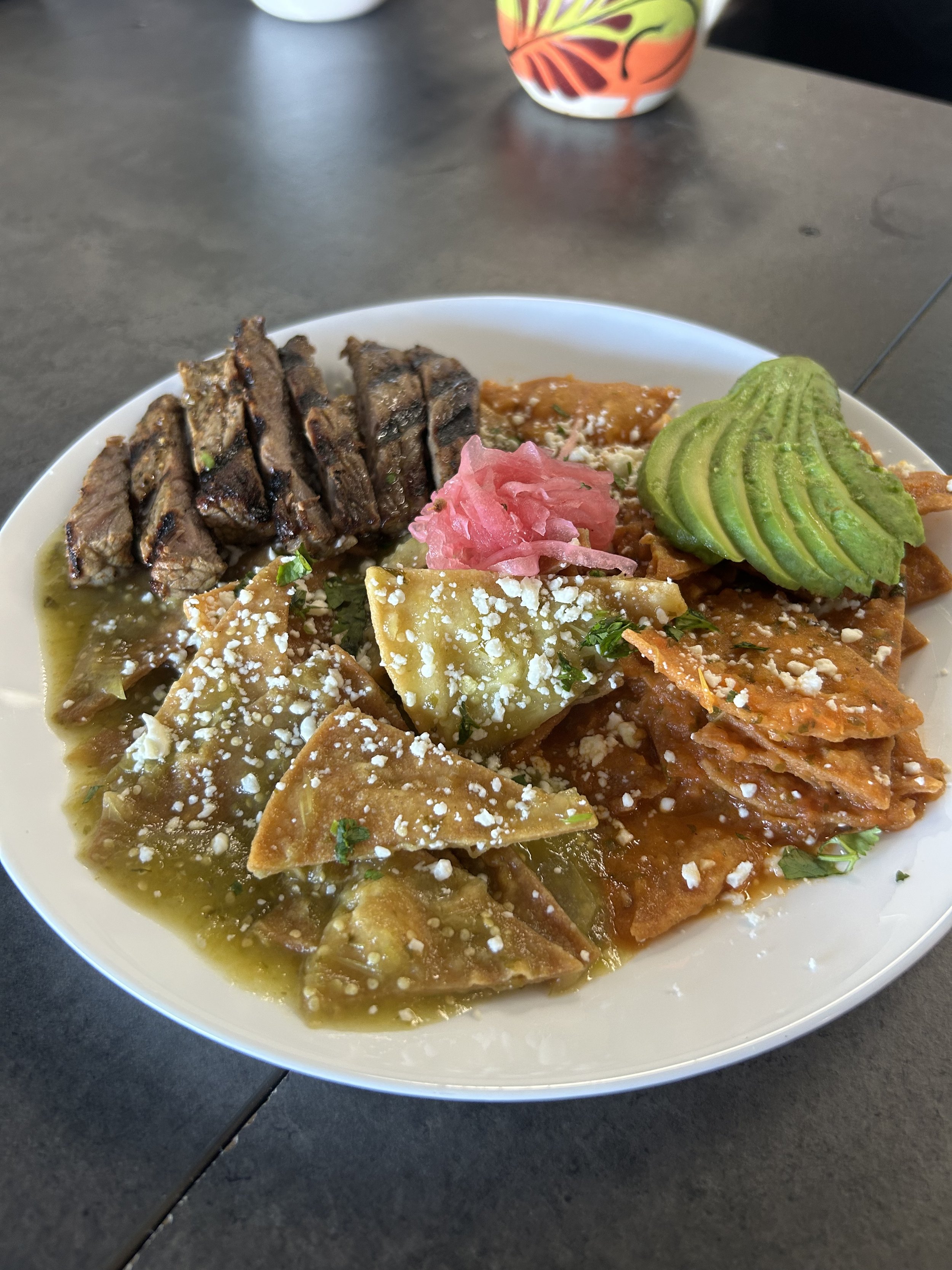 A white plate of assorted Mexican food including grilled meat, chicken with sauce and cheese, sliced avocado, pickled pink onions, and garnished with crumbled cheese and herbs.