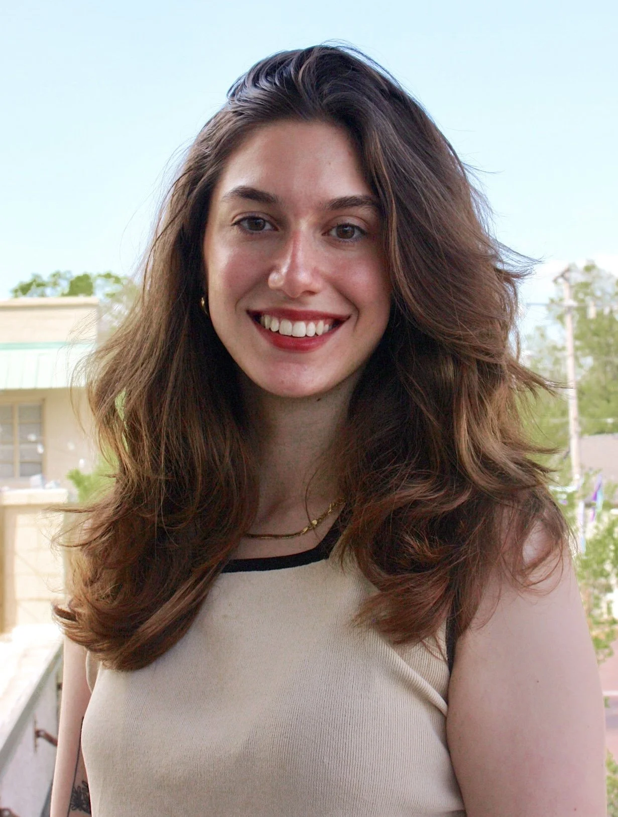 Woman with long brown hair smiling outdoors, wearing a light beige sleeveless top.