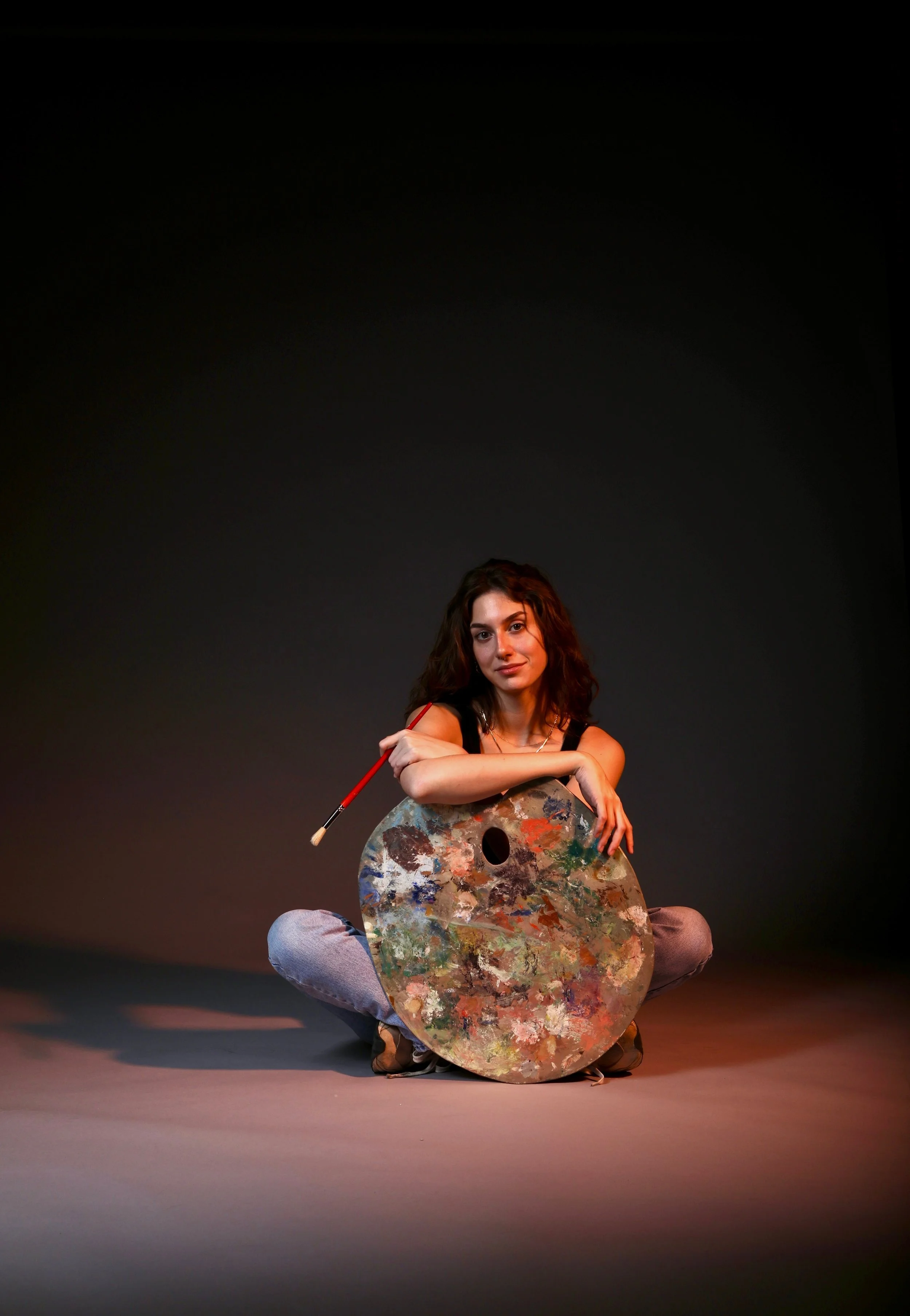 Artist sitting with a paintbrush and large paint palette in a studio setting.