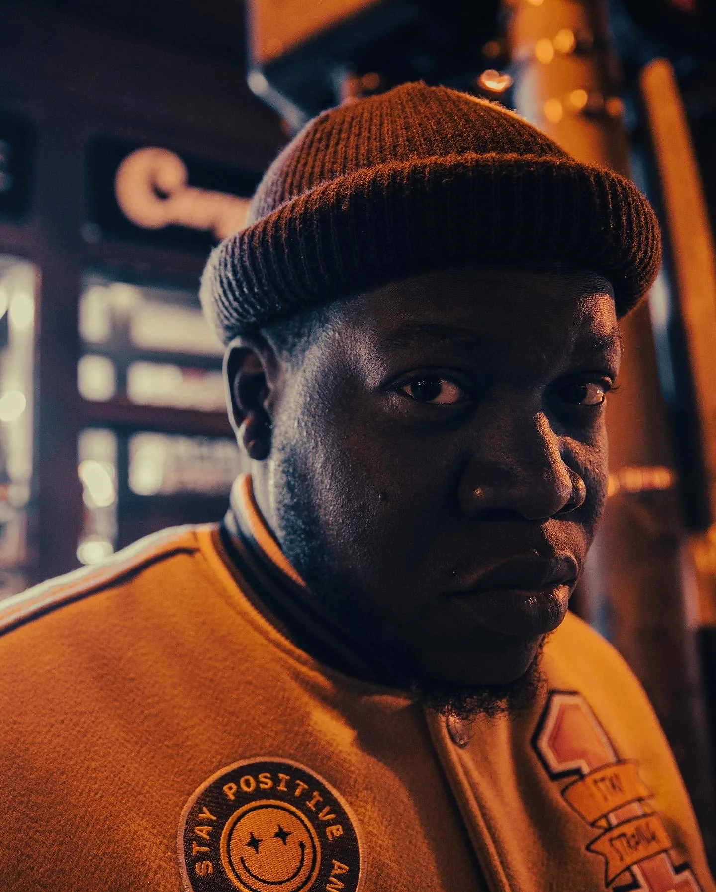 Person wearing a beanie and jacket with patches, standing at night on a city street.