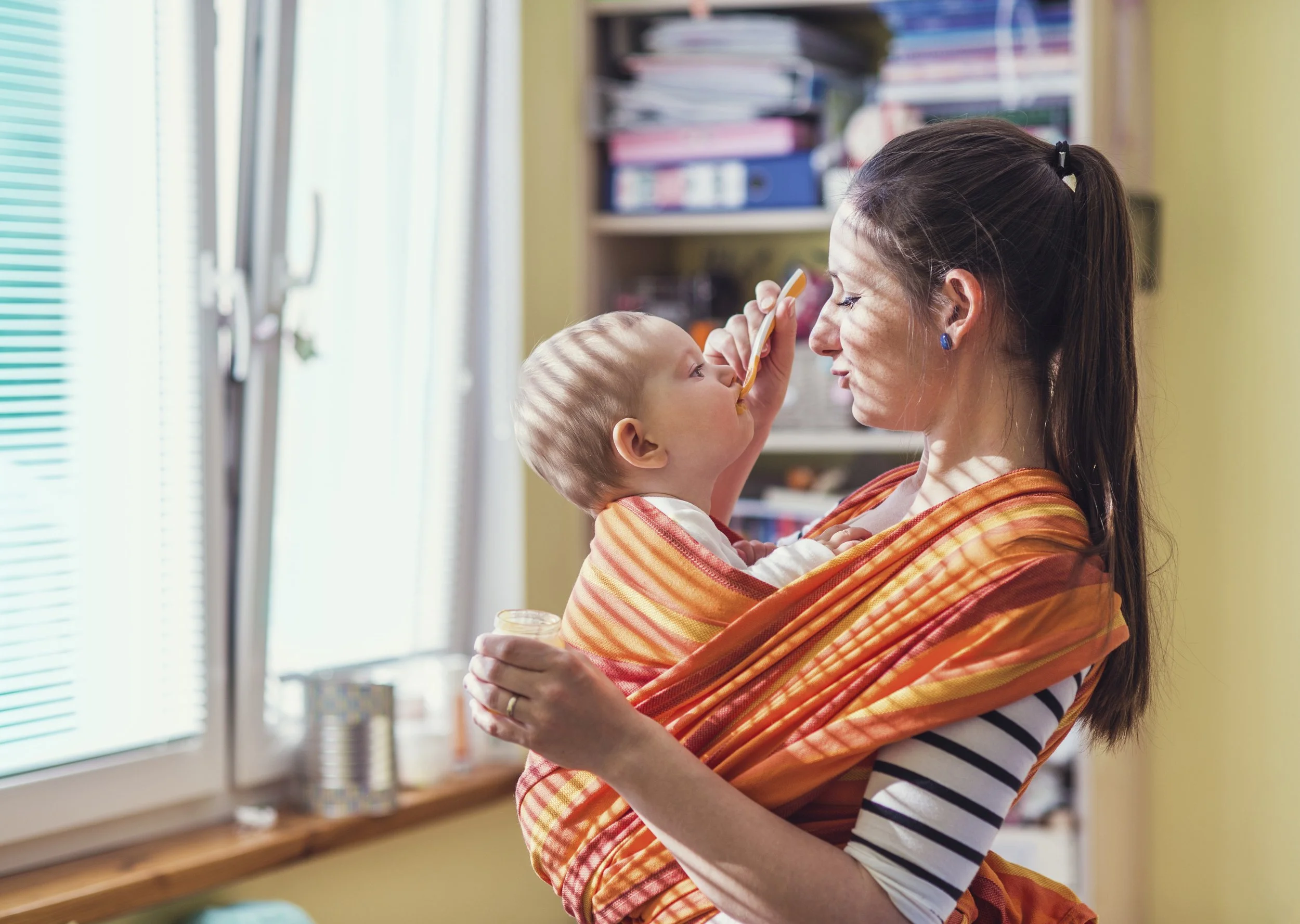 woman wearing her baby in an orange wrap and offering a spoon of solid foods