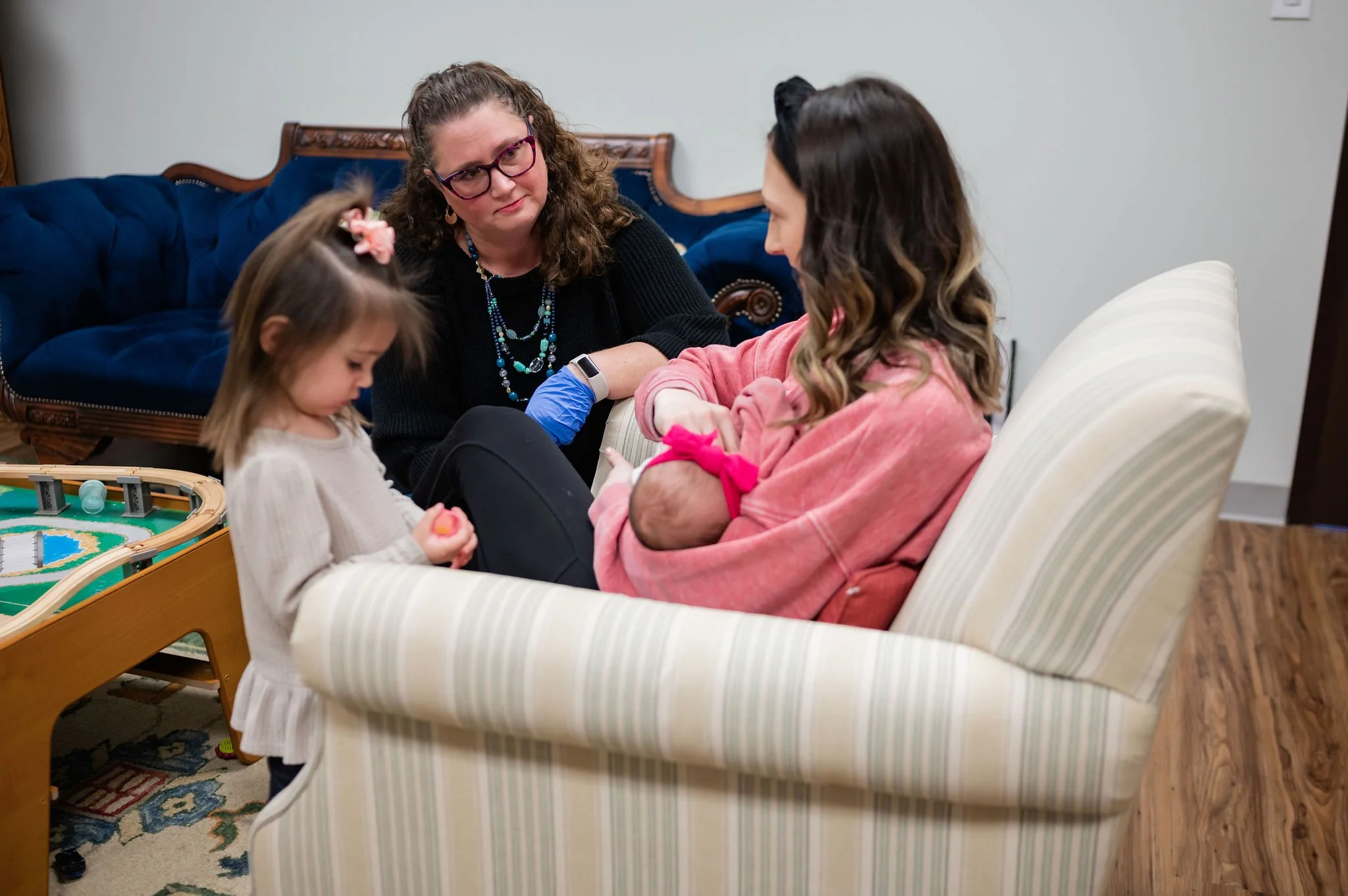 A woman in a pink sweatshirt is breastfeeding a baby while a young girl looks on and an older woman, possibly a nurse, observes.