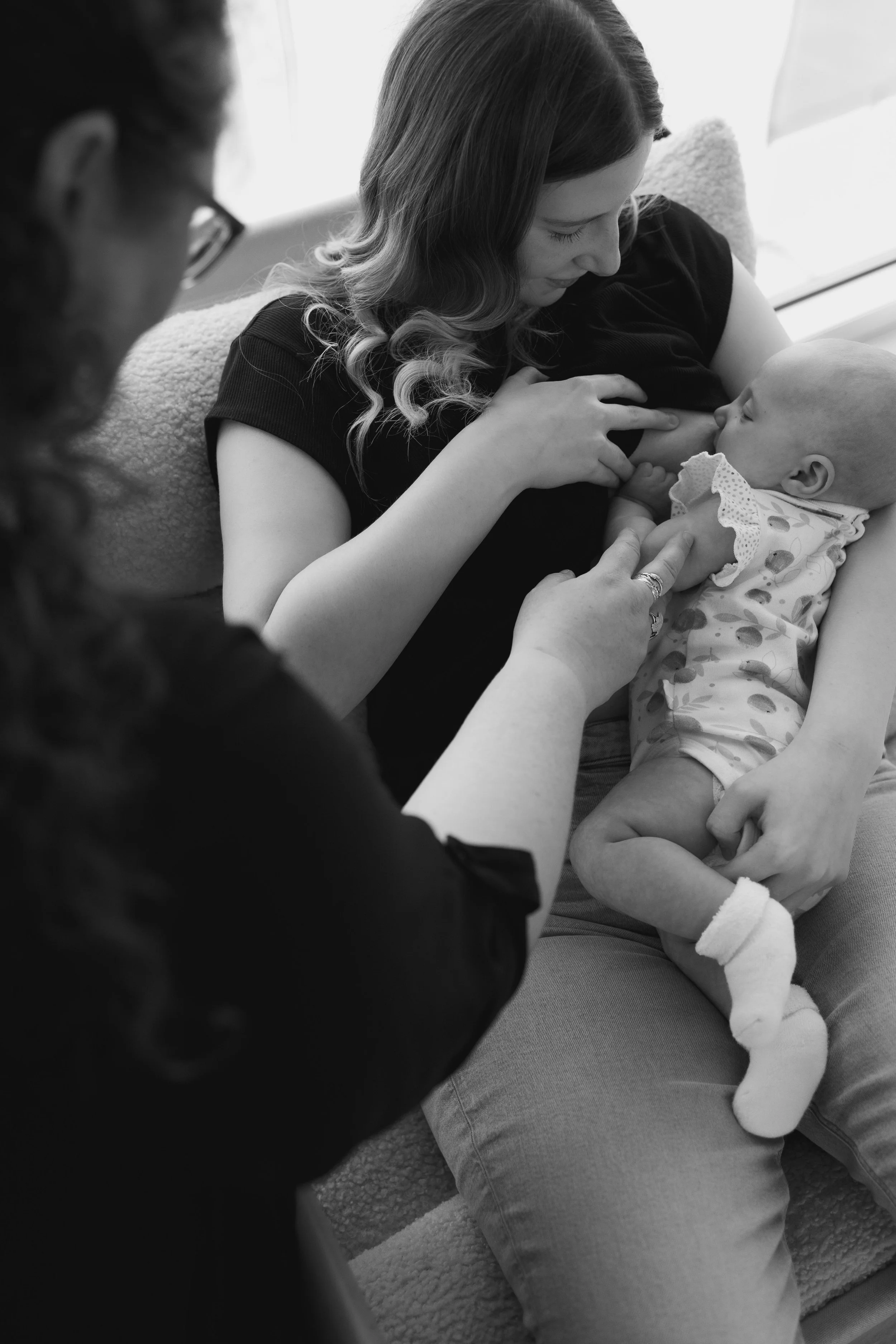 A woman with wavy hair breastfeeding an infant on her lap, while another woman assists and watches