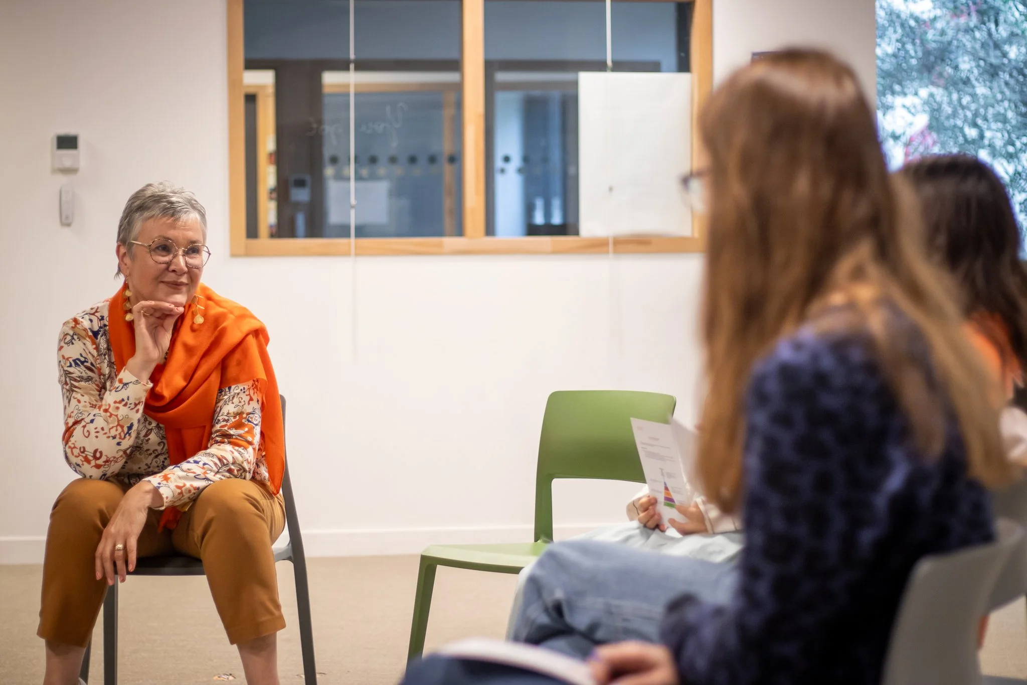 Corinne assise dans une pièce, semble donner une explication ou partager une expérience avec un groupe de personnes assises devant elle, dans un cadre de discussion ou de réunion.
