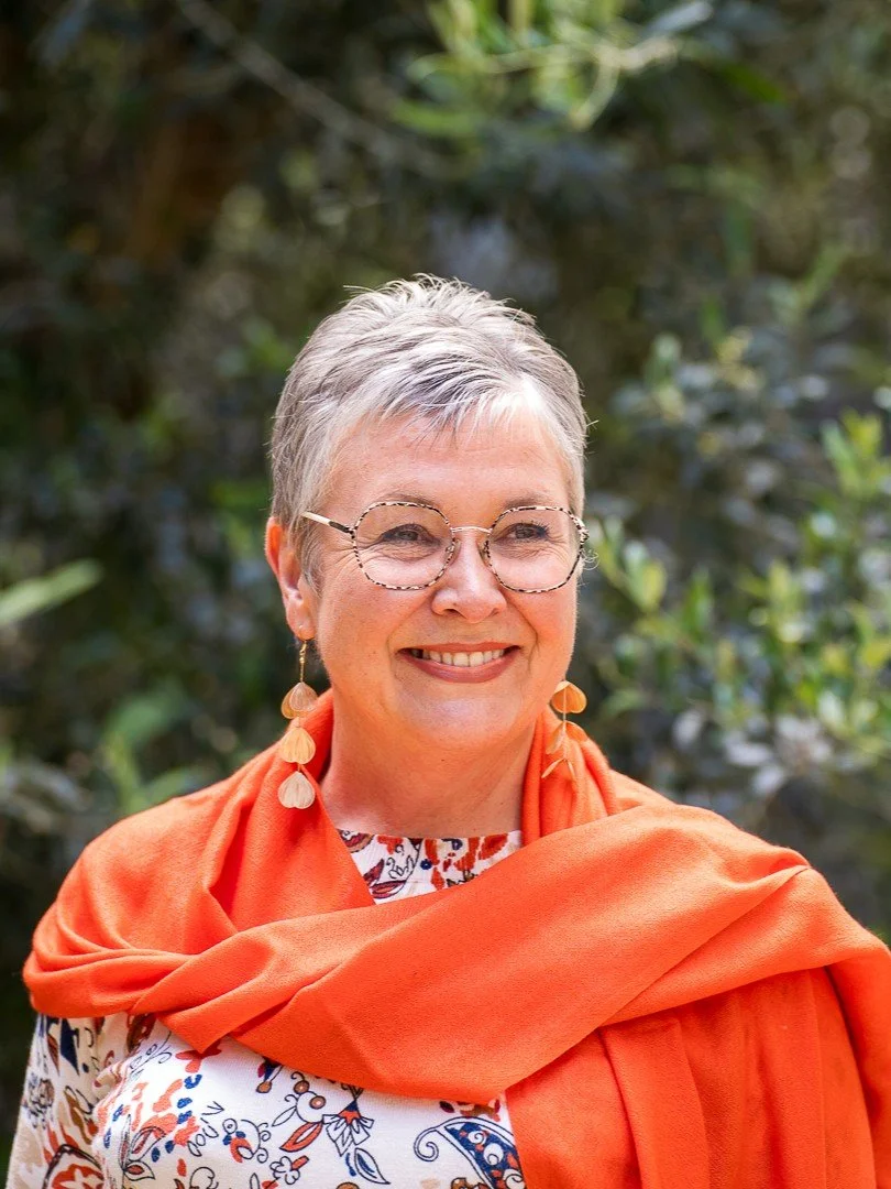 Corinne souriante, portant une robe avec un motif floral, une écharpe orange, et des boucles d'oreilles pendantes en forme de feuilles, en extérieur avec des buissons en arrière-plan.