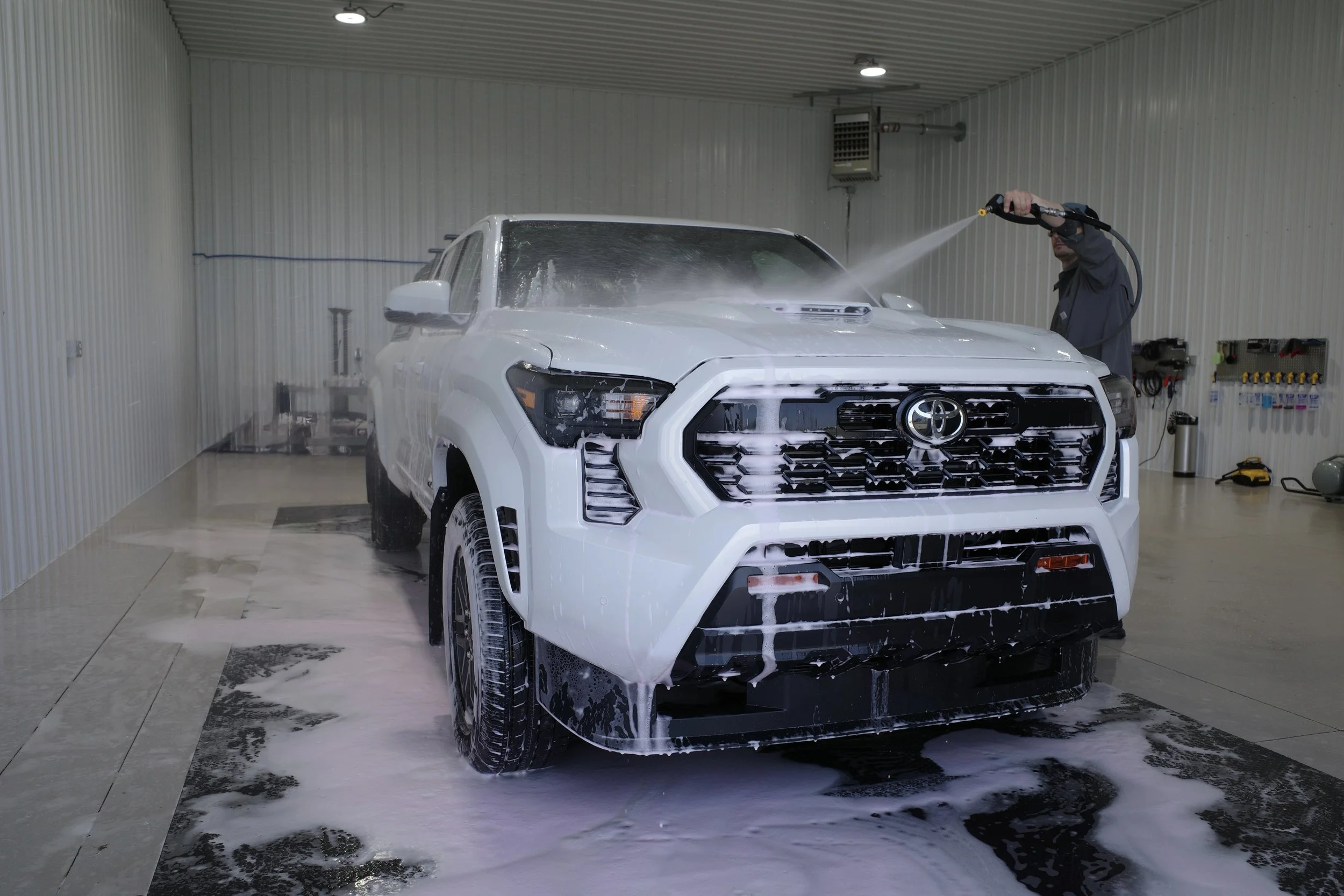 A white Toyota truck being washed with soap in an indoor car wash bay, using a high-pressure water hose.